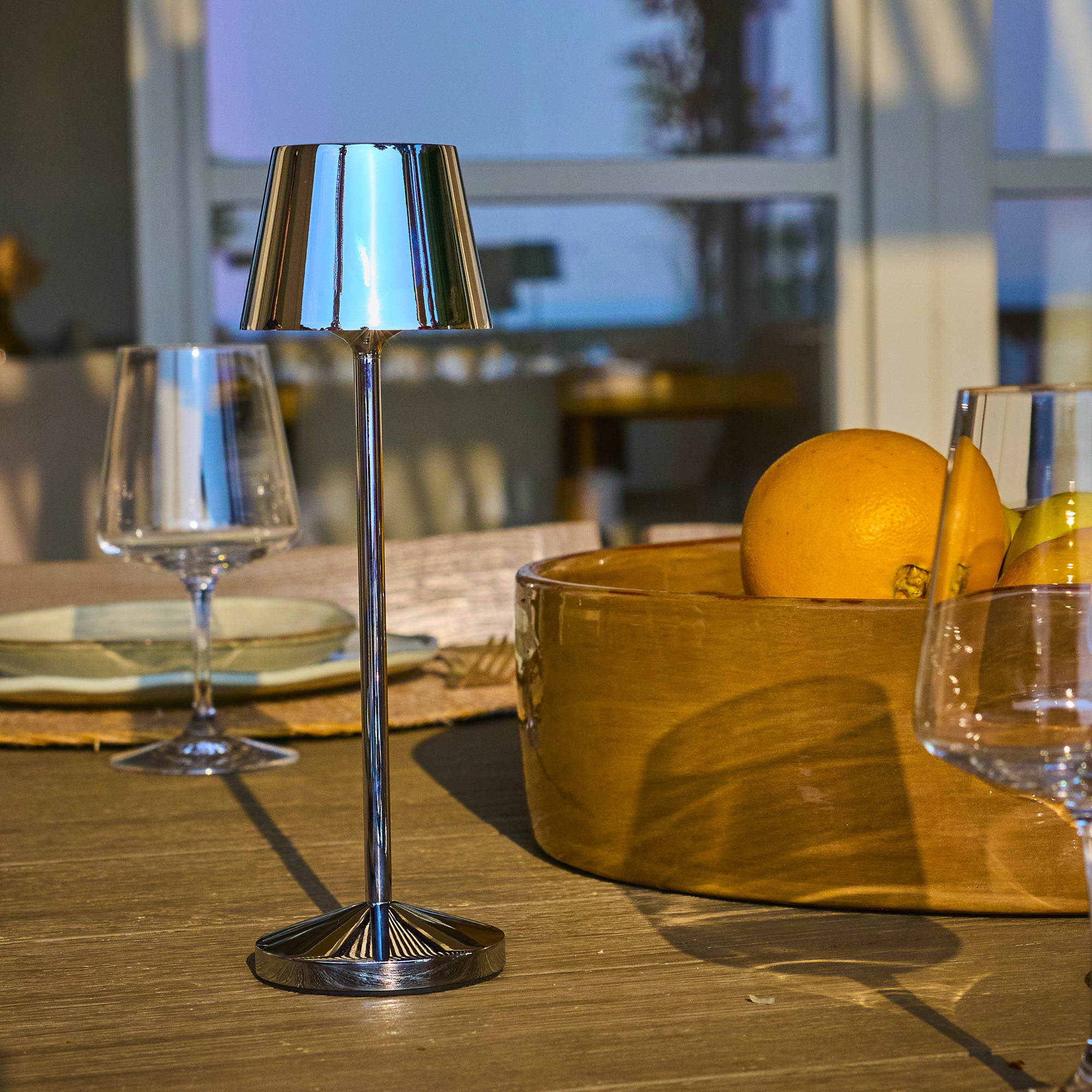 Tisch mit Lampe, Obstschale und Weingläsern. Sonnenlicht durchflutet den Hintergrund durch ein Fenster.
