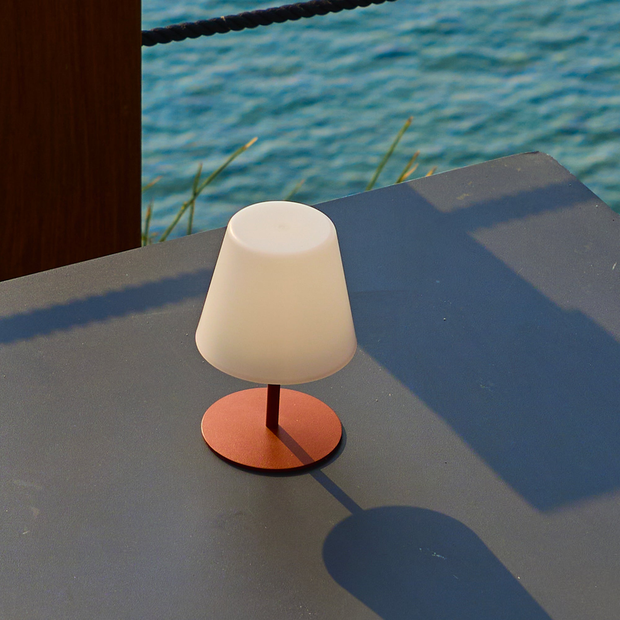 Eine kleine Lampe mit weißem Lampenschirm steht auf einem Tisch. Im Hintergrund sind Wasser und ein dunkler Holzpfosten zu sehen.