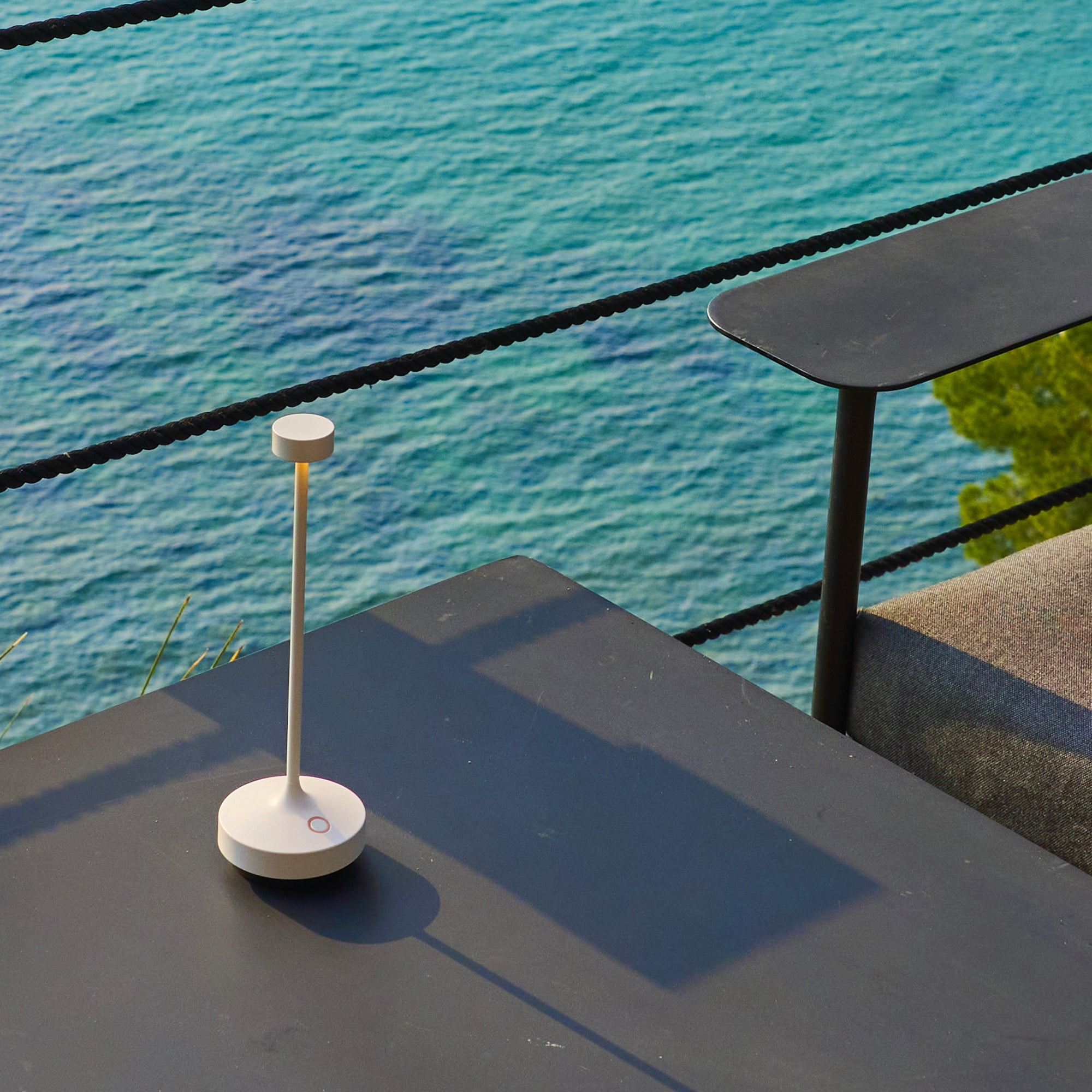 Eine moderne Lampe auf einem dunklen Tisch mit Blick auf türkisfarbenes Wasser und einen schwarzen Seilzaun.