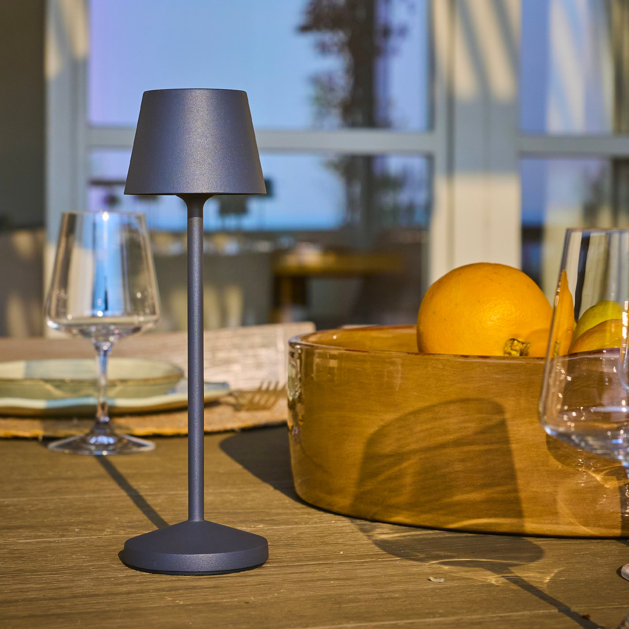 Tisch mit Lampe, Obstschale und Gläsern. Orangen und Zitronen sind sichtbar. Die Szene ist im Freien.