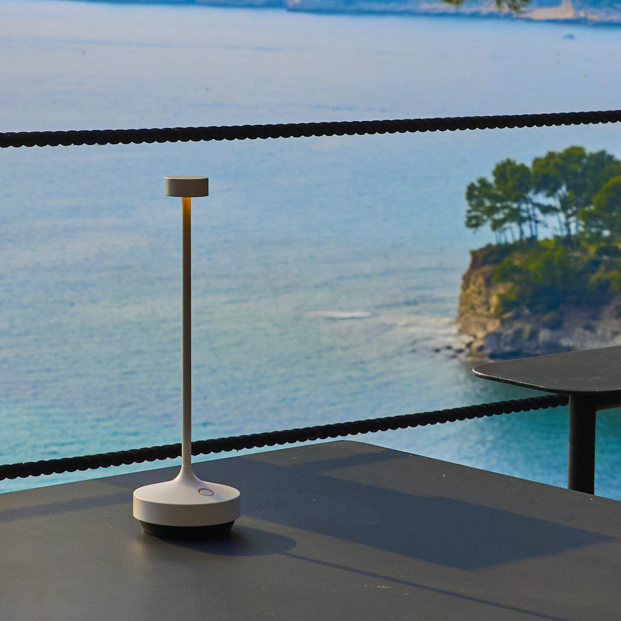 Eine weiße Lampe auf einem Tisch mit Meerblick. Eine kleine Insel mit Bäumen im Hintergrund. Schwarzer Seilzaun.