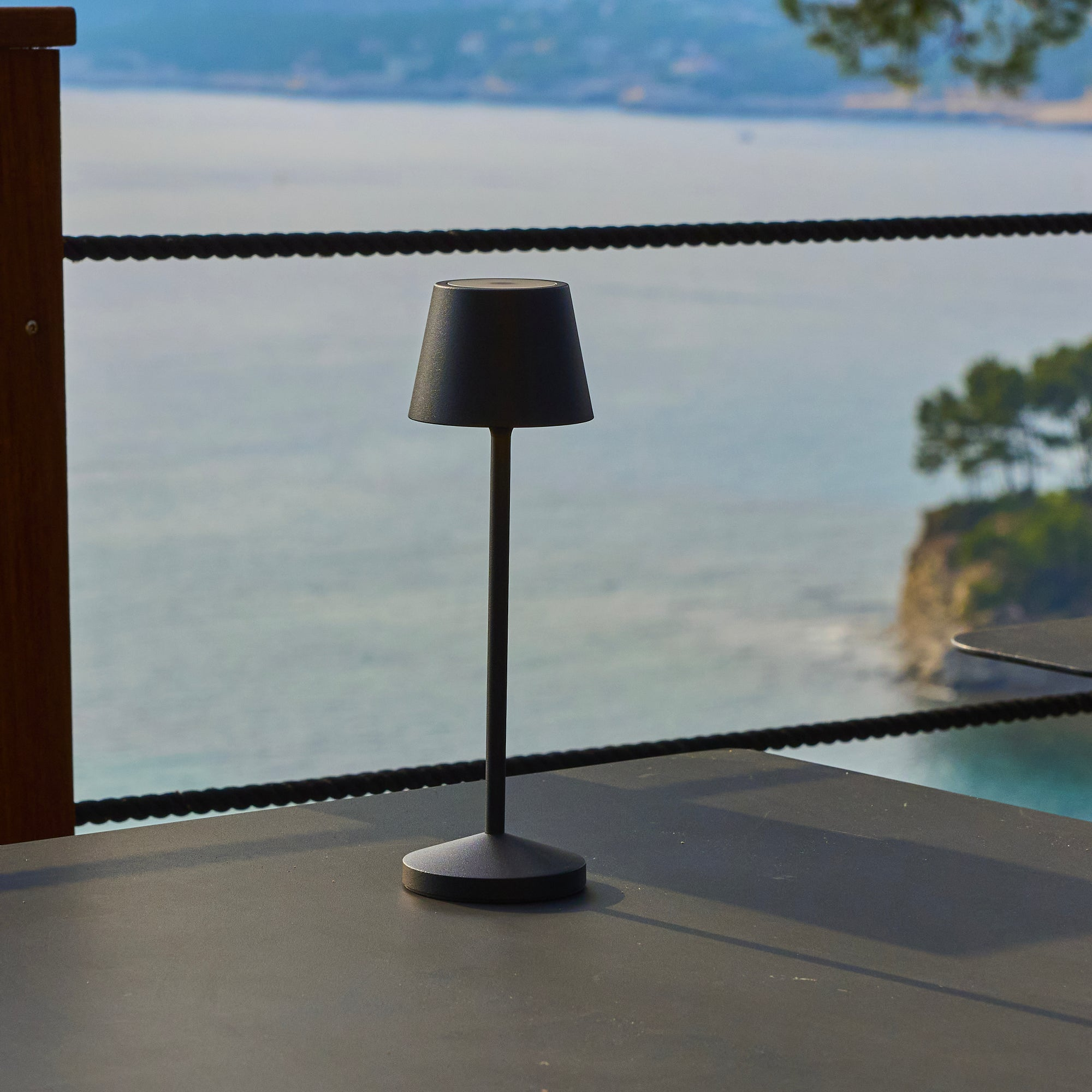 Eine schwarze Lampe steht auf einem Tisch. Im Hintergrund sind das Meer und eine Küstenlinie zu sehen. Ein Seilzaun verläuft über das Bild.