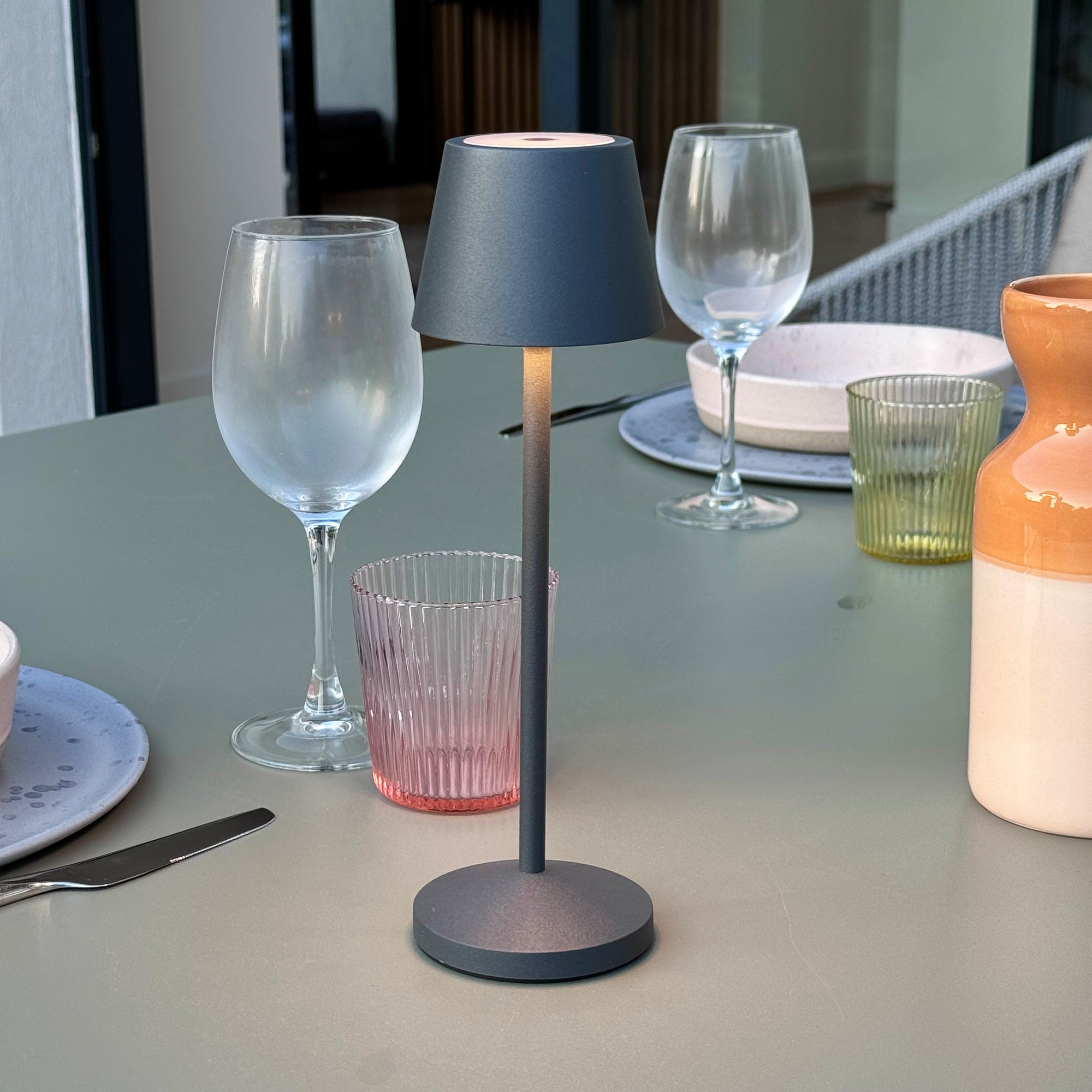 Tisch mit Gläsern, Tellern und einer Lampe. Ein rosa Glas und eine Vase sind ebenfalls sichtbar.