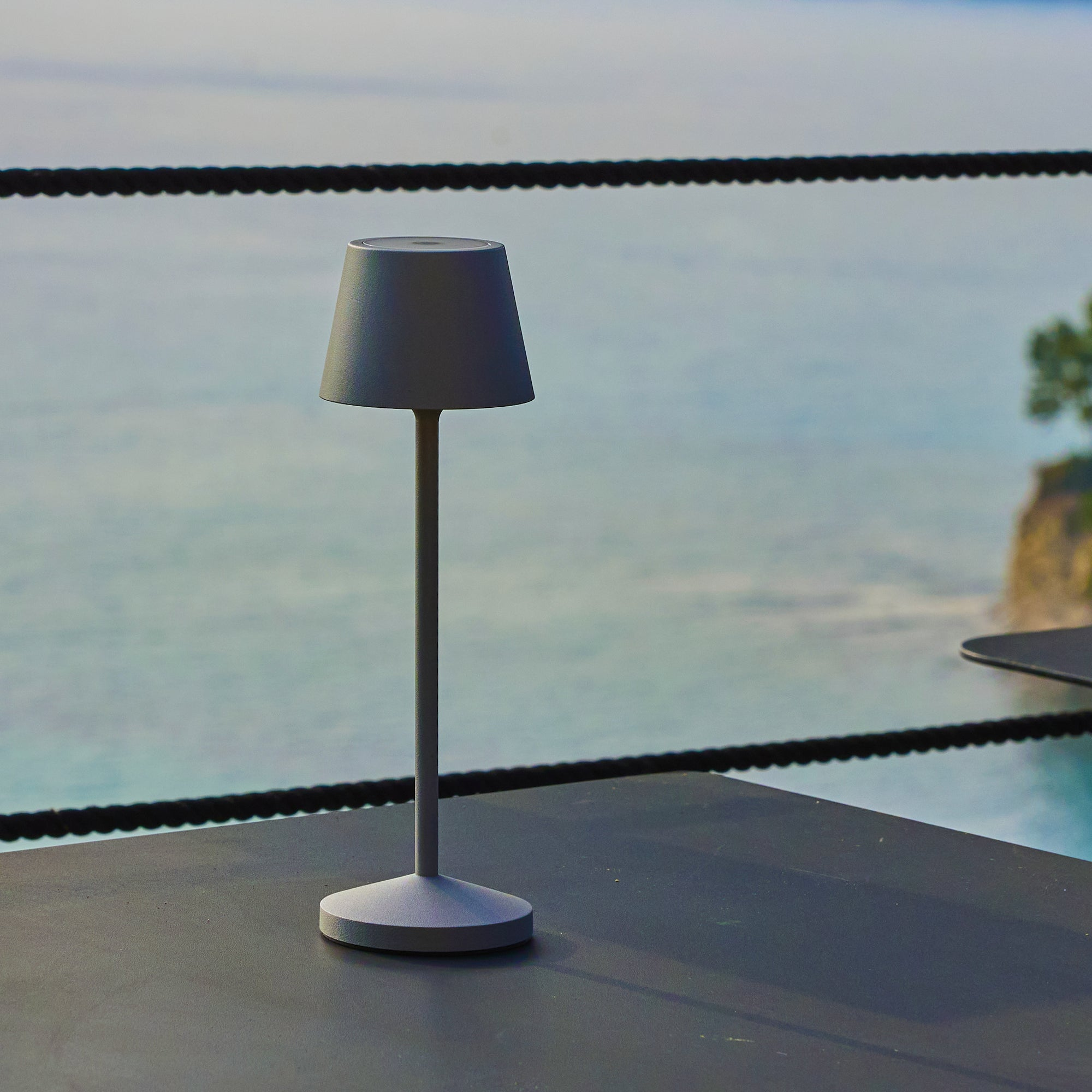 Eine graue Lampe auf einem Tisch, mit Meer und Himmel im Hintergrund. Ein schwarzes Seil befindet sich in der Mitte des Bildes.