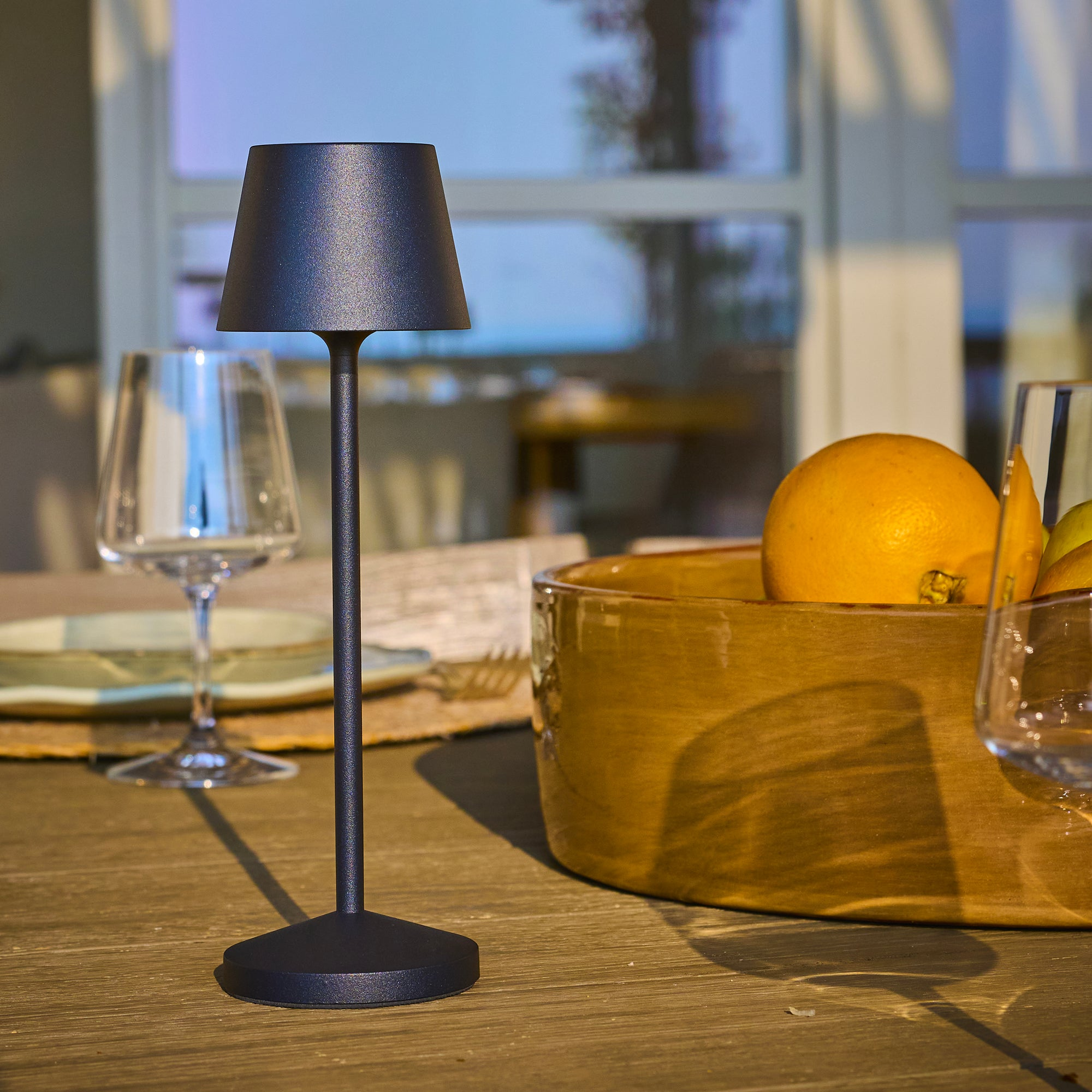 Eine dunkle Lampe steht auf einem Holztisch. Eine Schüssel mit Orangen und Gläsern stehen auf dem Tisch. Der Hintergrund ist unscharf.