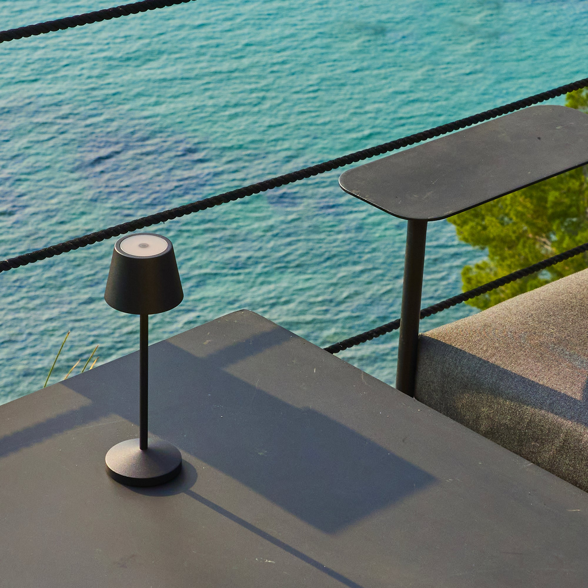 Eine schwarze Lampe auf einem Tisch mit Meerblick. Ein weiterer Tisch und ein Sofa sind sichtbar. Das Wasser ist türkis.
