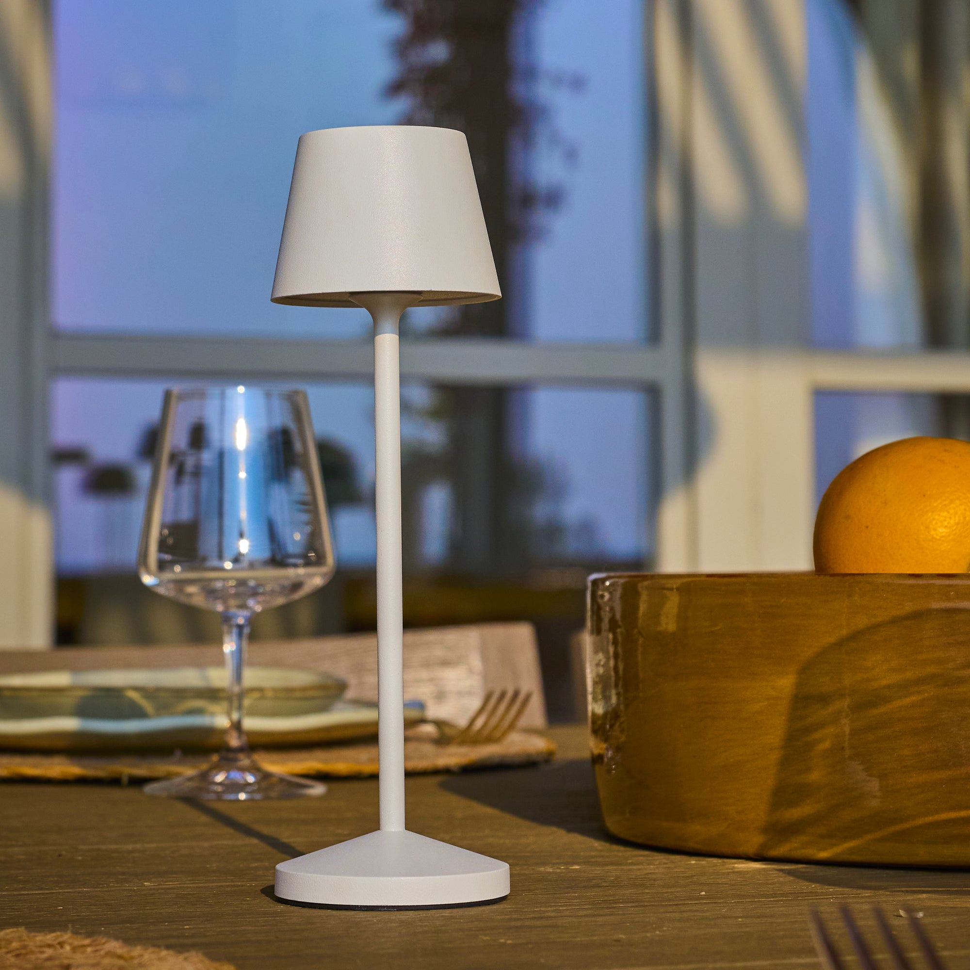 Eine weiße Lampe, ein Glas, eine Orange und eine Schüssel stehen auf einem Holztisch. Im Hintergrund ist ein Fenster.