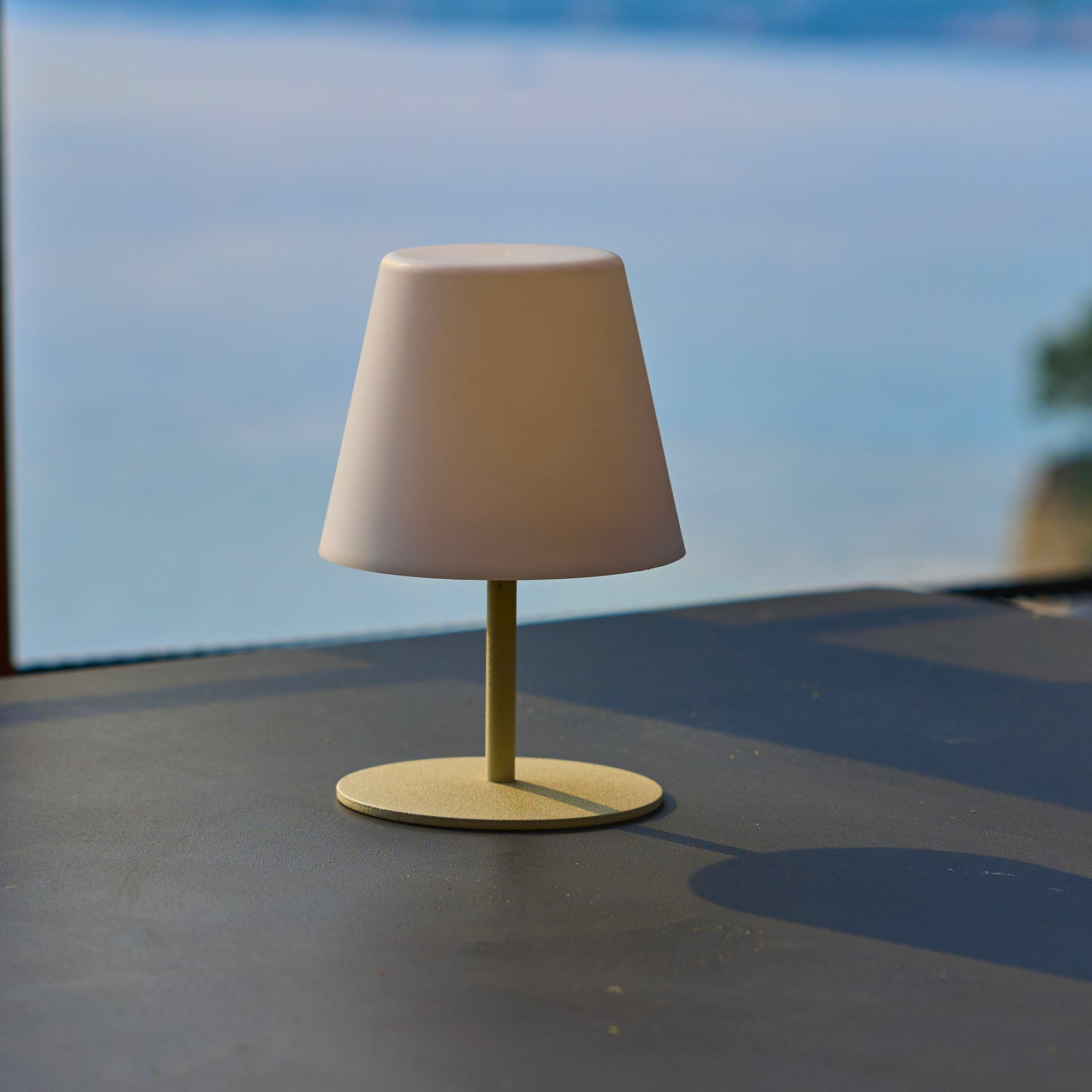 Eine kleine Lampe mit weißem Lampenschirm steht auf einem Tisch. Der Hintergrund zeigt das Meer und einen blauen Himmel.