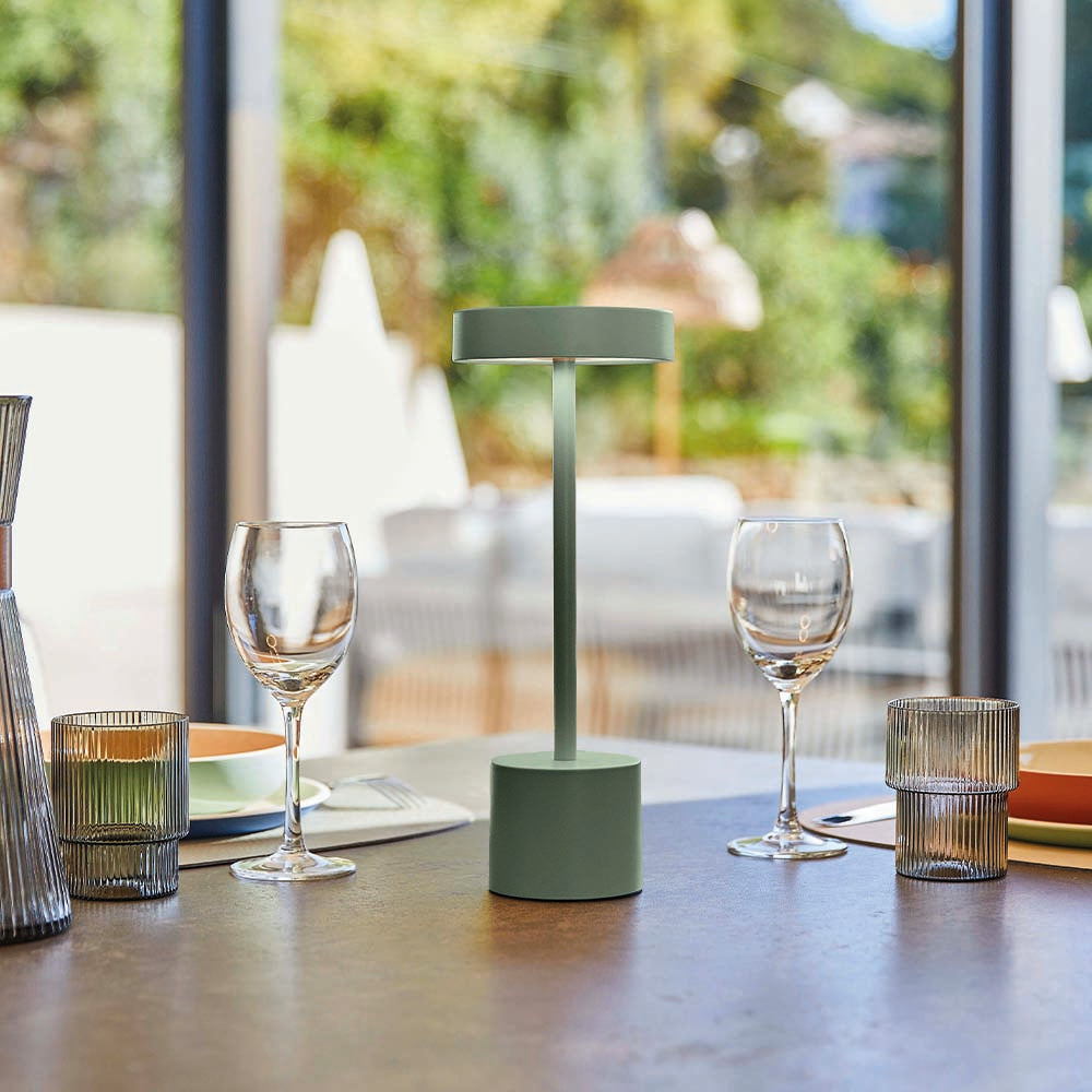 Eine grüne Lampe steht auf einem Tisch. Weingläser und andere Glaswaren sind vorhanden. Der Hintergrund zeigt ein Fenster mit Bäumen.
