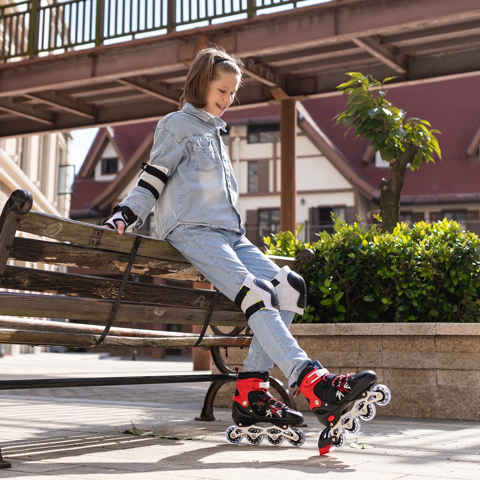 Una niña en mezclilla con patines se sienta en un banco. Edificios y vegetación están al fondo.