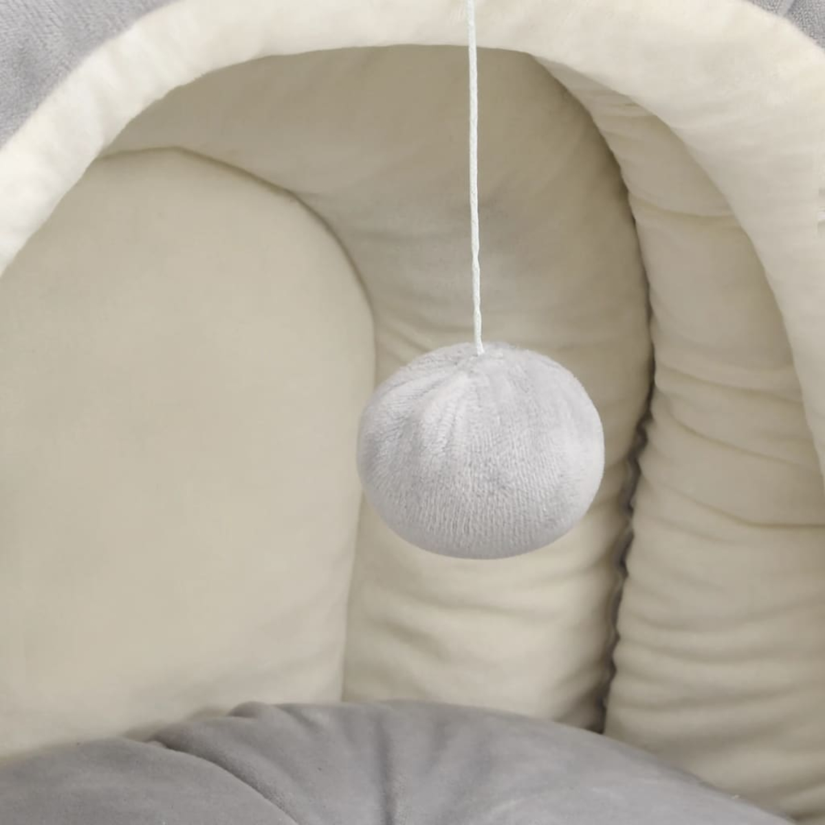 Ein graues Katzenbett mit einem hängenden, flauschigen, grauen Ball. Das Bett ist innen weiß, außen grau.
