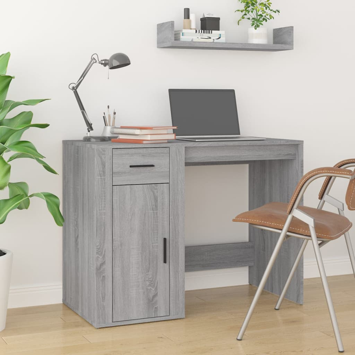 Ein grauer Schreibtisch mit Laptop, Lampe und Büchern. Daneben steht ein brauner Stuhl. Im Vordergrund eine Pflanze, im Hintergrund ein Regal.