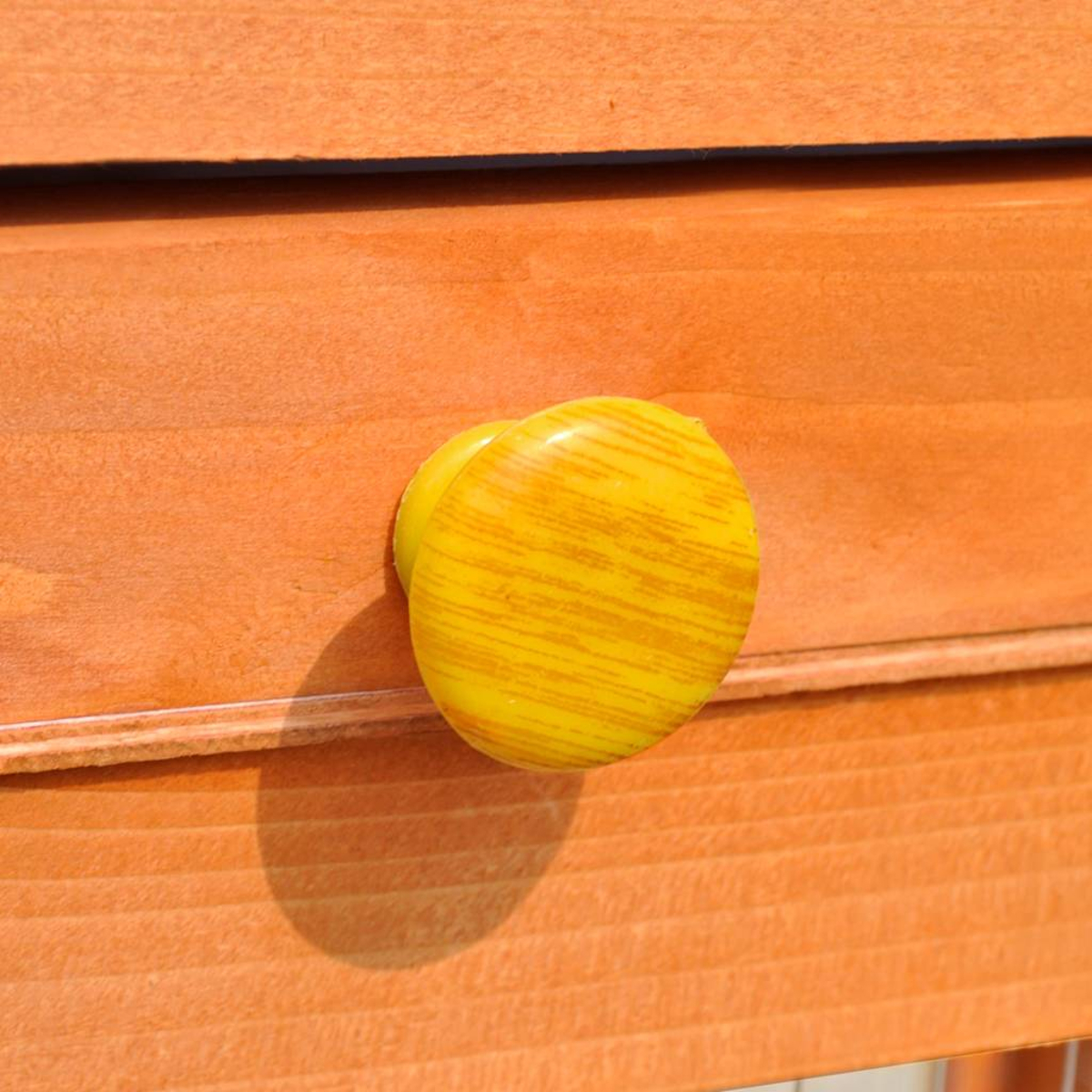 Nahaufnahme zeigt gelben, gestreiften Knopf auf orangefarbener Holzoberfläche. Der Knopf wirft einen Schatten. Die Holzmaserung ist sichtbar.