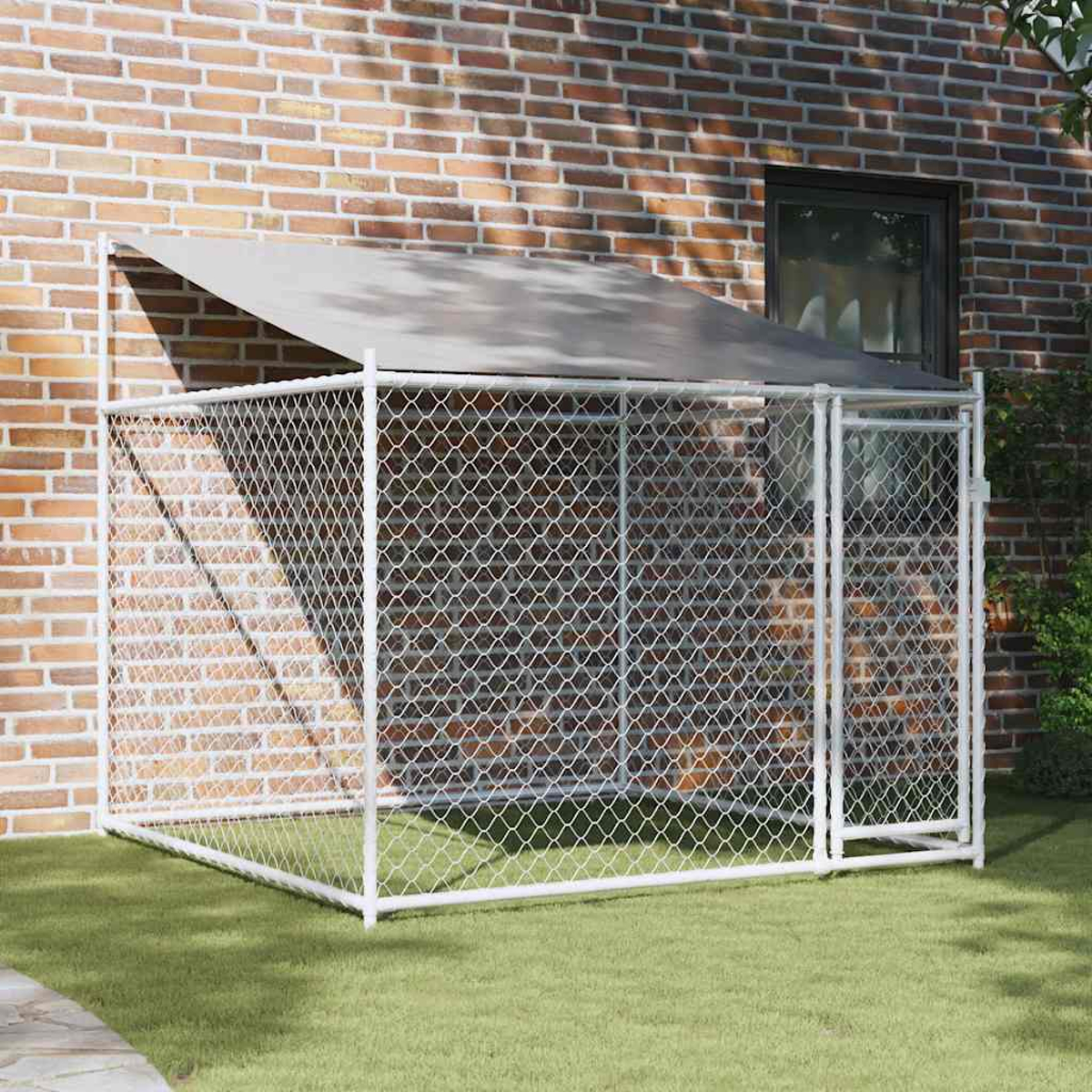 Ein weißer Zwinger mit Maschendrahtzaun steht auf grünem Gras. Eine Backsteinmauer und ein Fenster sind im Hintergrund.