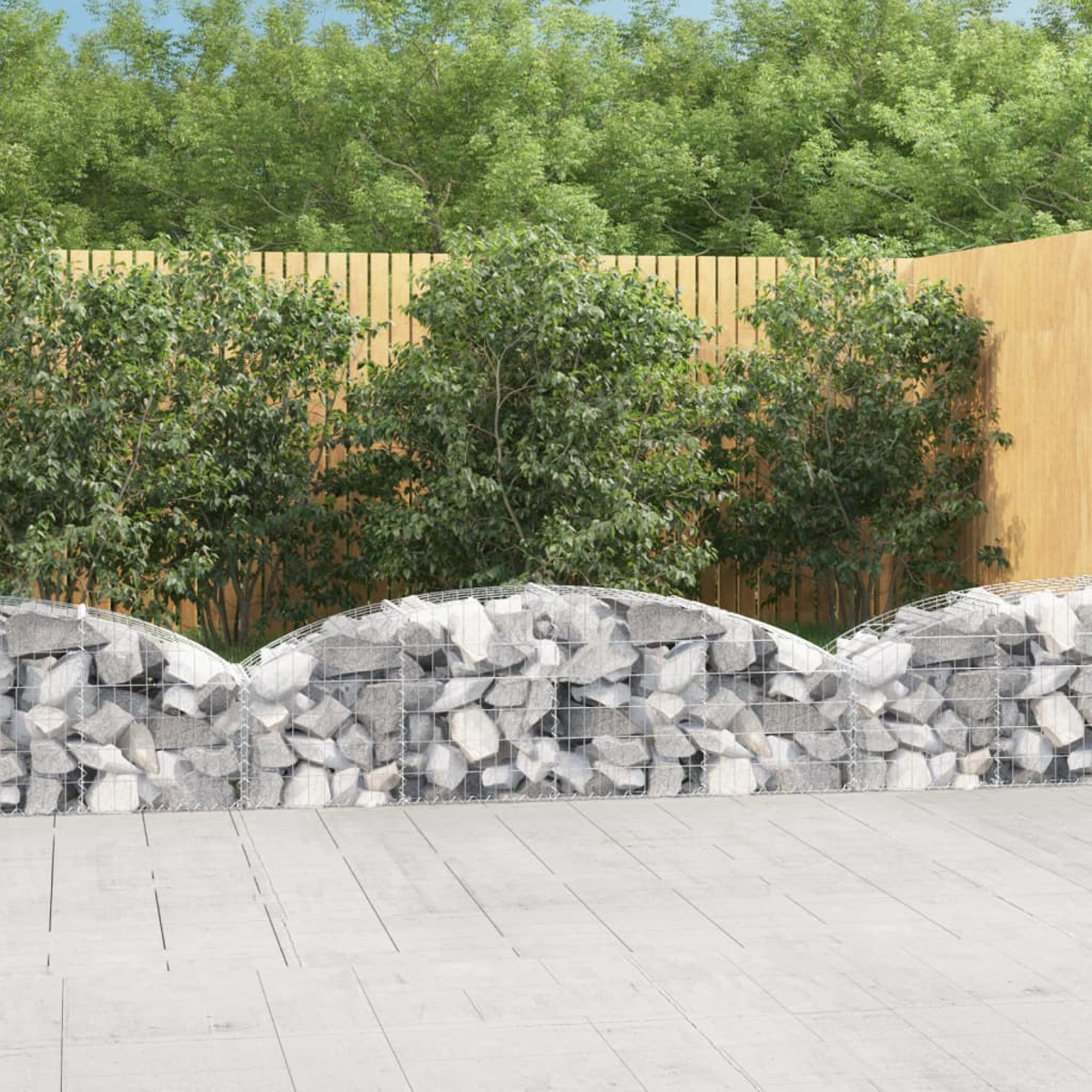 Eine Steinmauer mit Drahtgeflecht, dahinter grüne Büsche, ein Holzzaun und Bäume. Im Vordergrund befindet sich eine Beton-Terrasse.