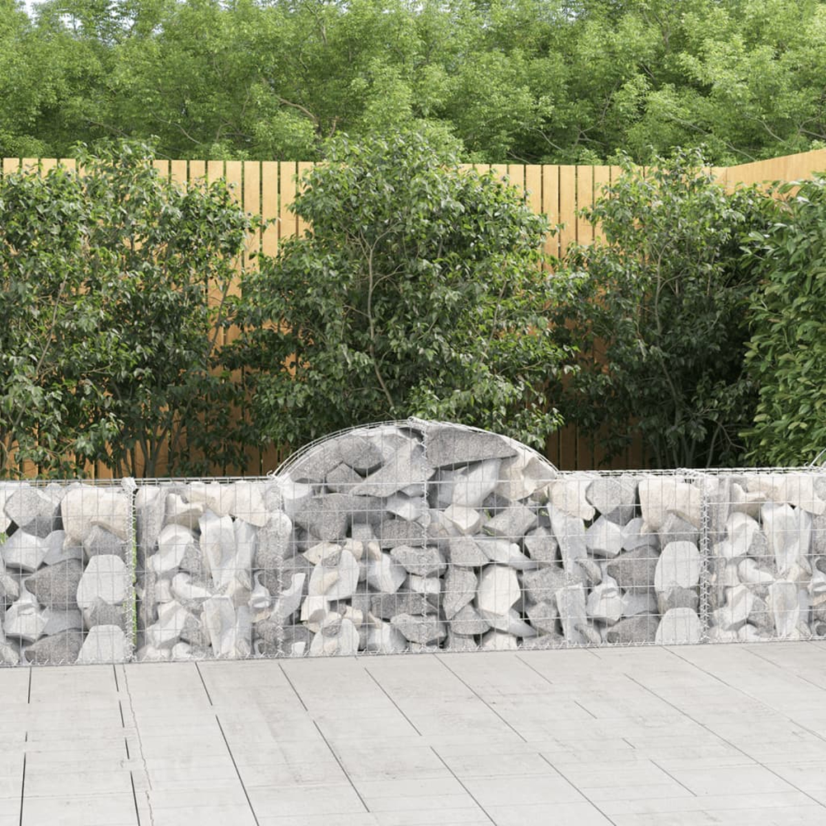 Eine Steinmauer mit Drahtrahmen. Dahinter befinden sich grüne Büsche und ein Holzzaun. Der Boden besteht aus hellgrauem Beton.