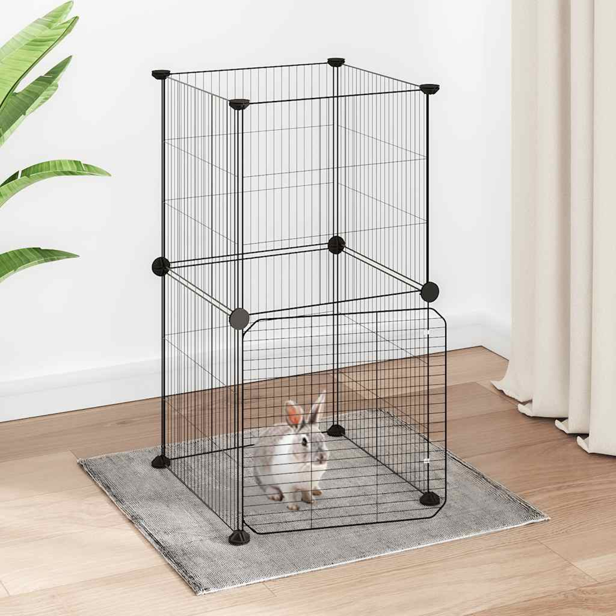 Ein weiß-graues Kaninchen sitzt in einem schwarzen Drahtkäfig auf einem grauen Teppich. Der Käfig ist zweistöckig.