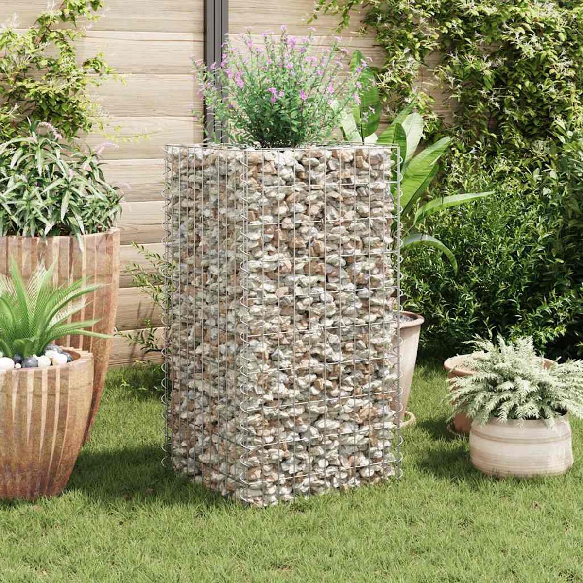 Ein mit Steinen gefüllter Gabion steht in einem Garten. Pflanzen in Töpfen und üppiges Grün umgeben ihn. Im Hintergrund befindet sich ein Holzzaun.