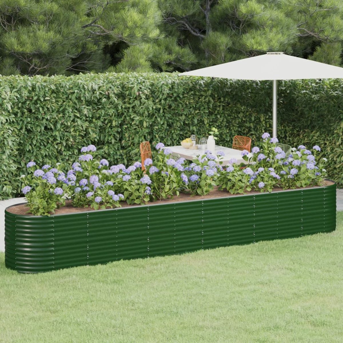 Ein grünes Hochbeet mit lila Blumen, einem Tisch und Stühlen unter einem Sonnenschirm, vor einer grünen Hecke.