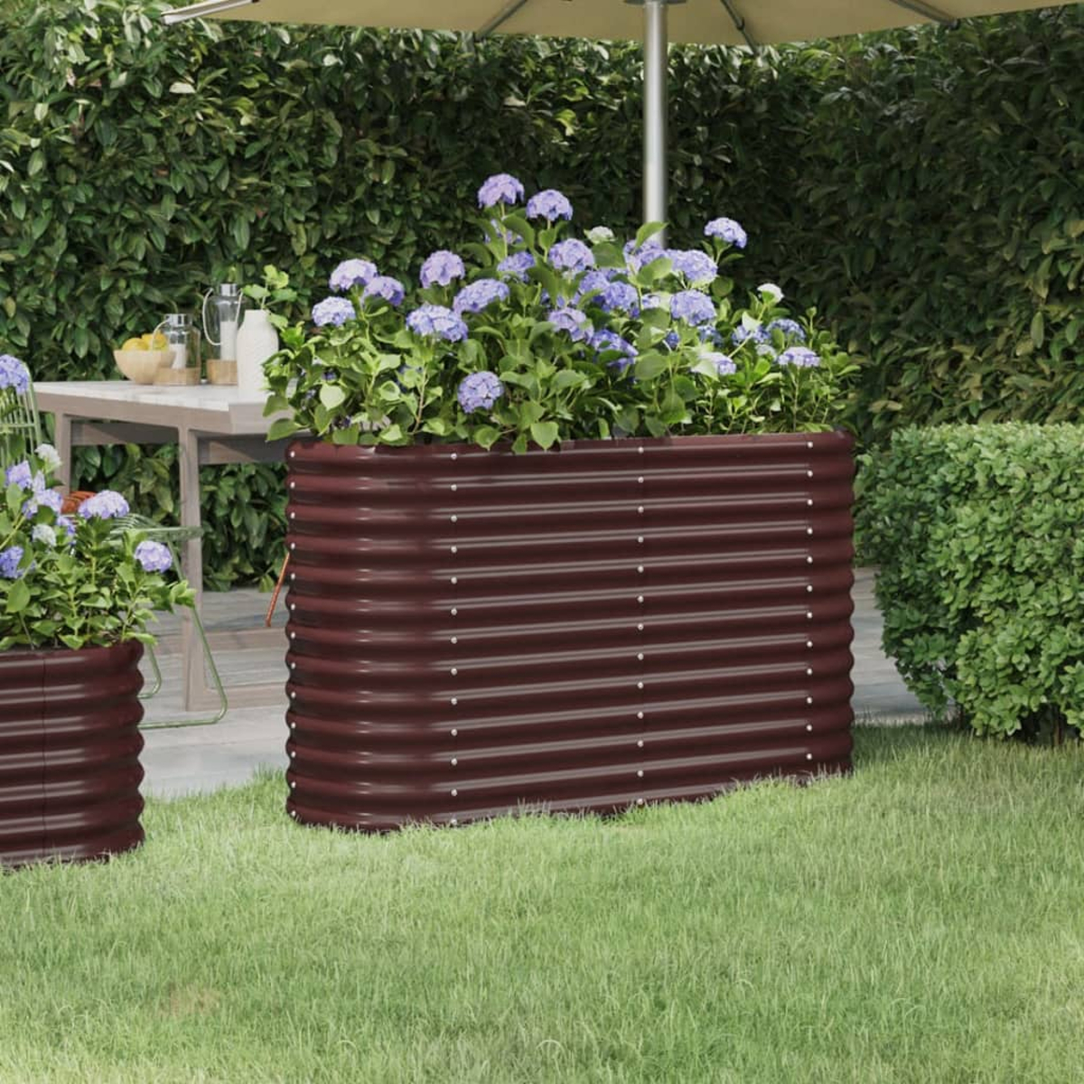 Gartenszene mit großem, weinrotem Metallpflanzgefäß mit blauen Blumen. Im Hintergrund ein Tisch und ein Sonnenschirm.