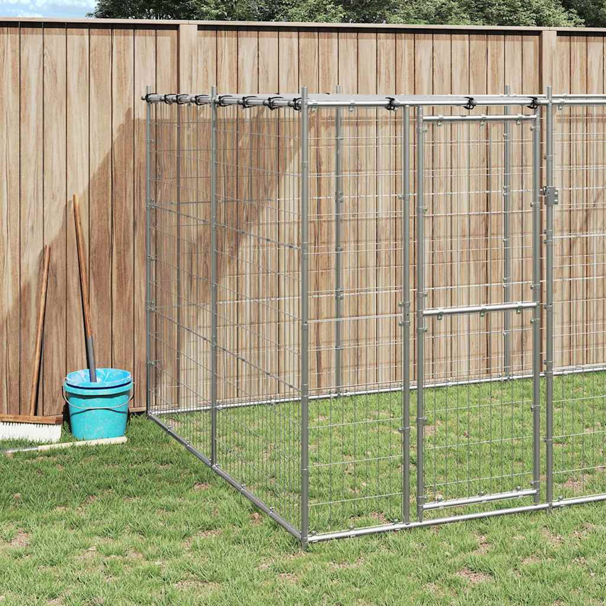Metall-Hundegehege auf grünem Gras, neben einem Holzzaun. Ein blauer Eimer und Reinigungswerkzeuge stehen neben dem Zaun.