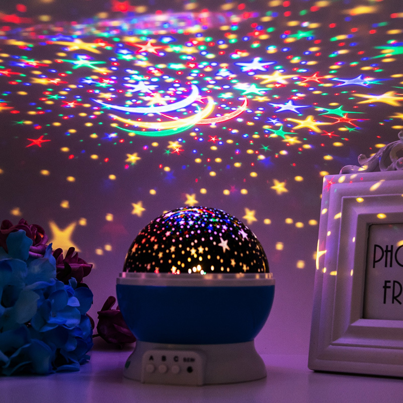 Ein Sternprojektor projiziert bunte Sterne und Halbmonde auf eine lila Wand. Eine blaue Lampe steht im Vordergrund mit Blumen und einem Bilderrahmen.