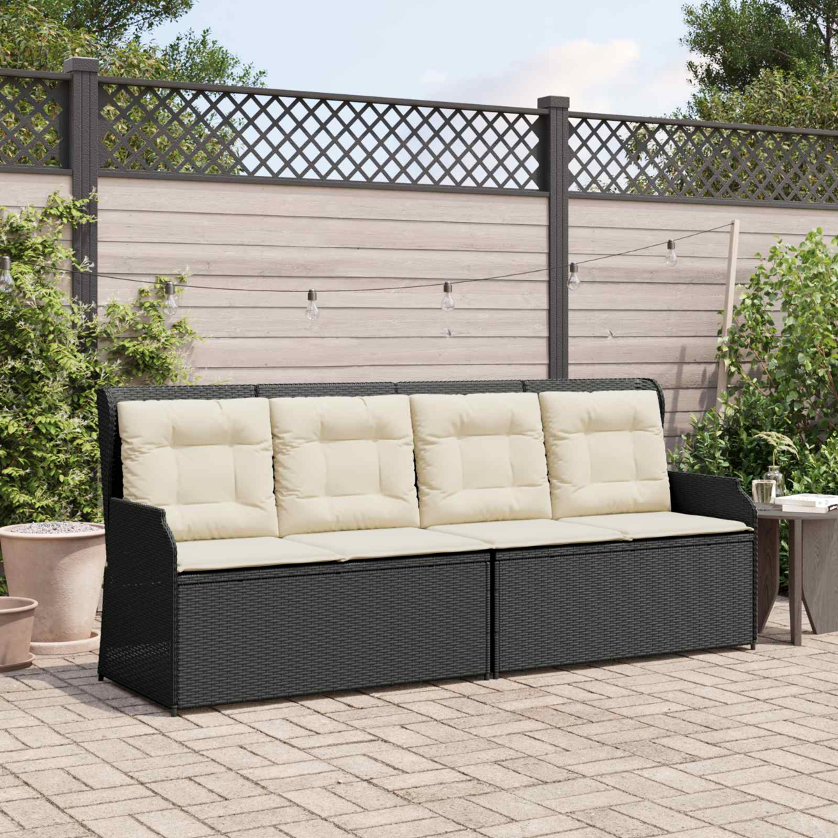Ein dunkelgraues Korbmöbelsofa mit cremefarbenen Kissen steht auf Ziegeln. Im Hintergrund sind Holzzaun und Pflanzen zu sehen.