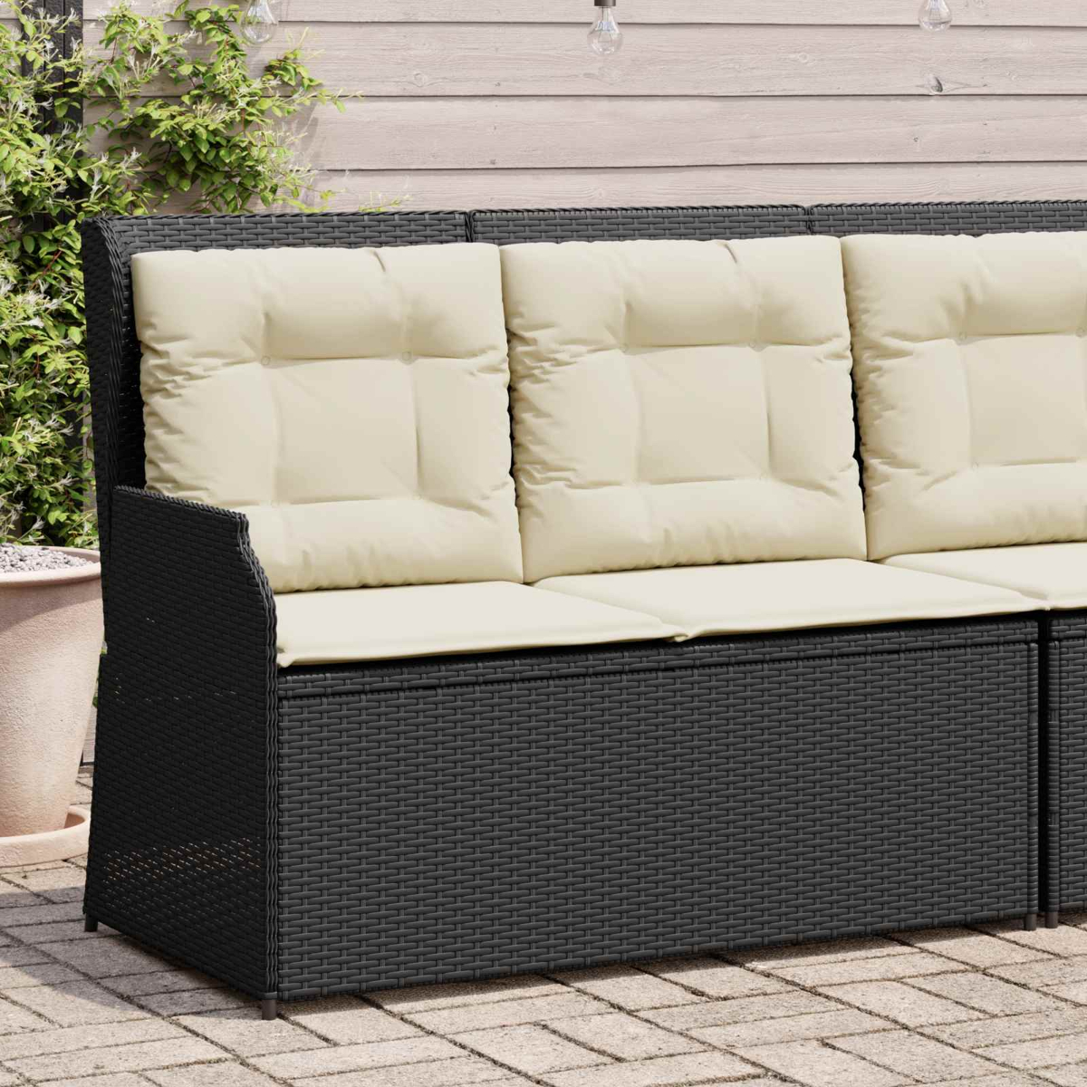 Schwarzes Outdoor-Sofa mit cremefarbenen Kissen. Im Hintergrund eine Holzwand, Pflanzen und Lichterketten.