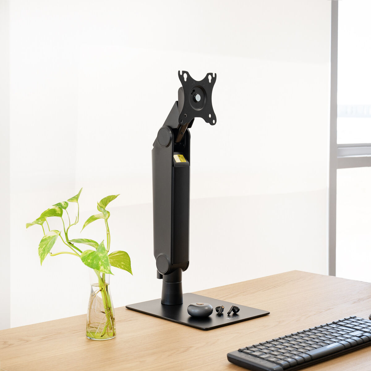 Un braccio nero per monitor su una scrivania di legno. Una piccola pianta in un vaso di vetro a sinistra. Una tastiera in basso a destra.