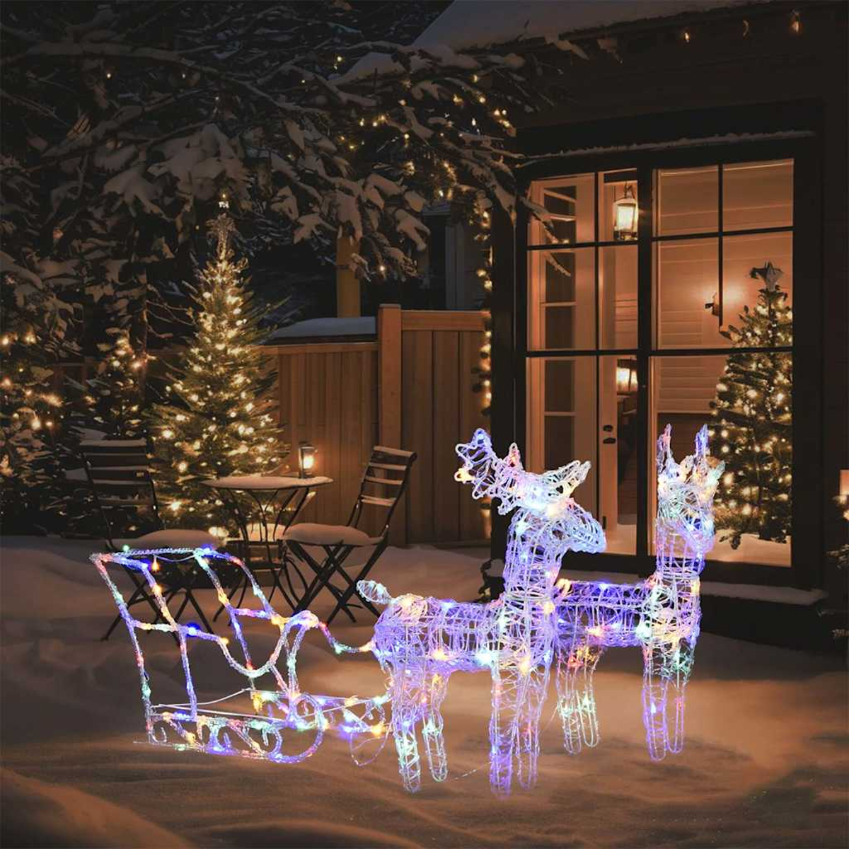 Beleuchtete Rentier- und Schlittendekoration in einem verschneiten Garten. Ein Haus mit Weihnachtsbeleuchtung in den Fenstern. Bäume und Schnee im Hintergrund.