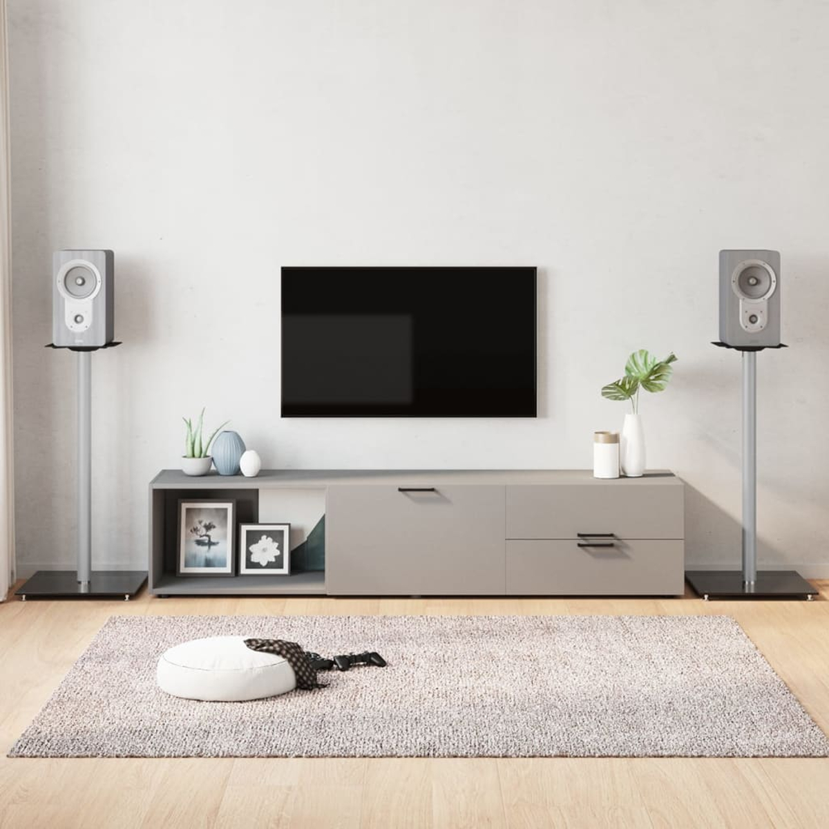 Wohnzimmer mit Fernseher, Lautsprechern und einem grauen Schrank. Ein Teppich mit einem weißen Pouf und einem gemusterten Schal liegt auf dem Boden.