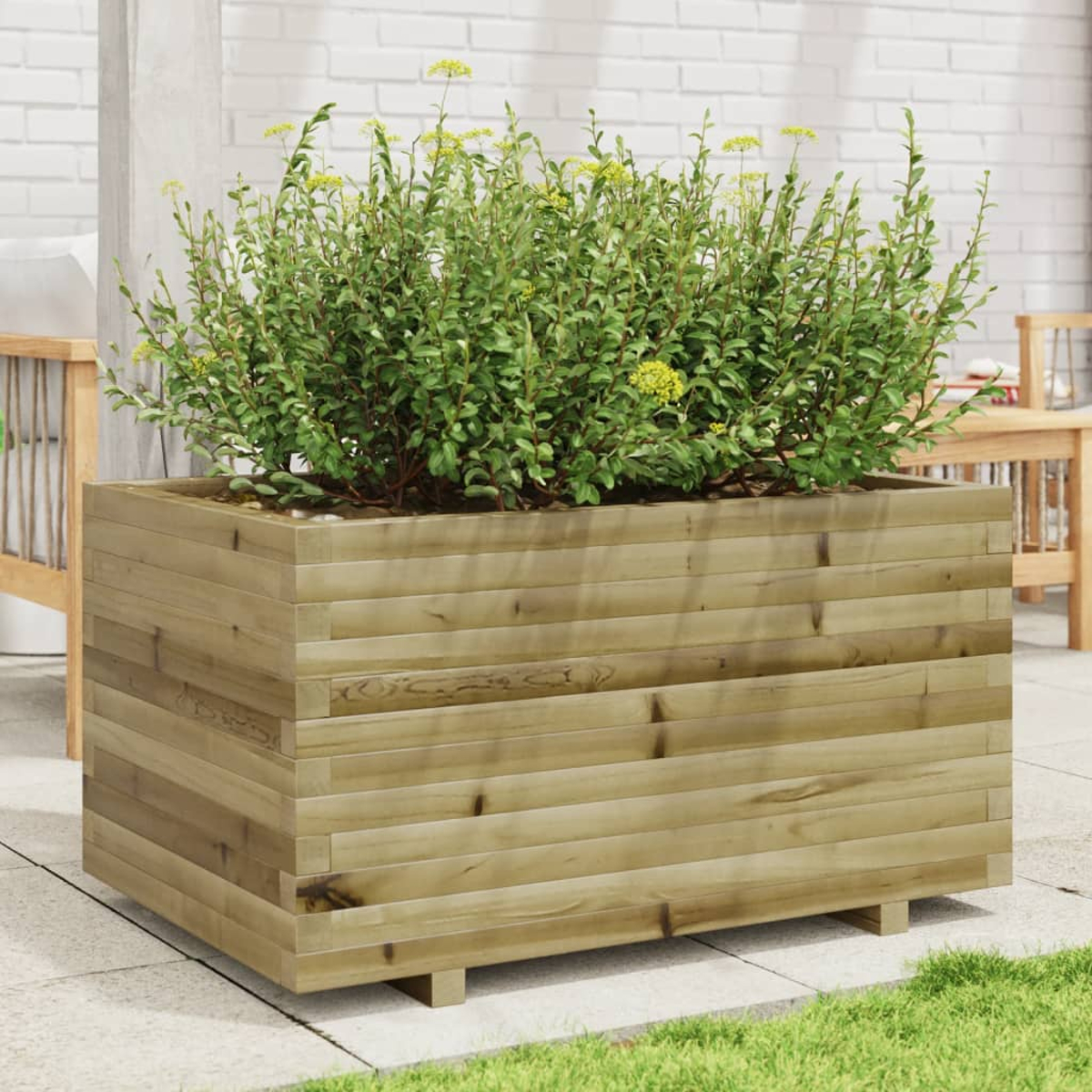 Eine große Holzkiste mit grünen Pflanzen und gelben Blüten. Im Hintergrund eine Terrasse mit Möbeln und einer weißen Backsteinmauer.