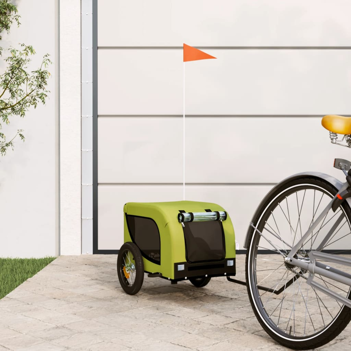 Ein grüner Fahrradanhänger mit einer orangefarbenen Fahne, die an einem Fahrrad befestigt ist. Das Fahrrad steht auf einem gepflasterten Bereich.