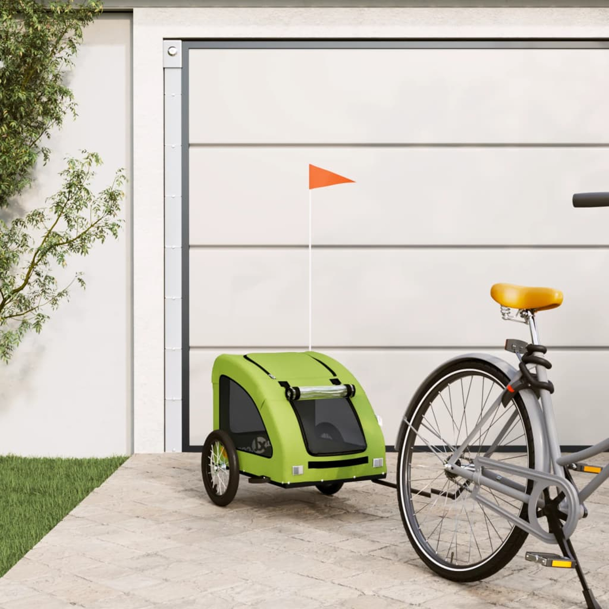 Ein grüner Fahrradanhänger mit orangefarbener Fahne ist an einem grauen Fahrrad befestigt. Der Hintergrund hat ein Garagentor und einen Baum.