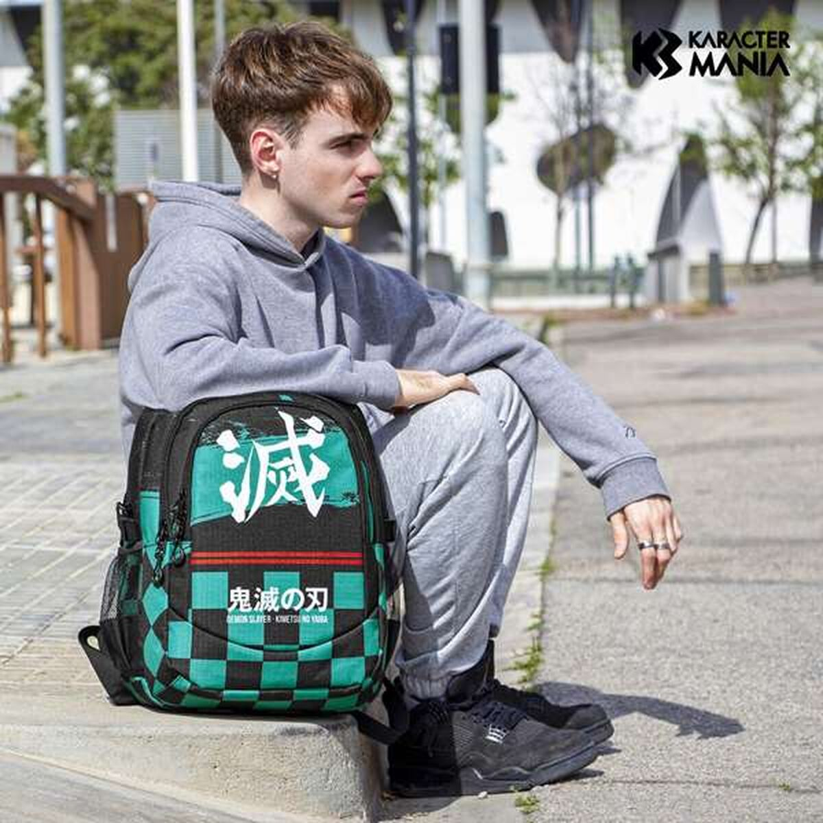Ein junger Mann sitzt neben einem Rucksack. Der Rucksack ist schwarz, grün und weiß. Er trägt einen grauen Hoodie und eine Jogginghose.