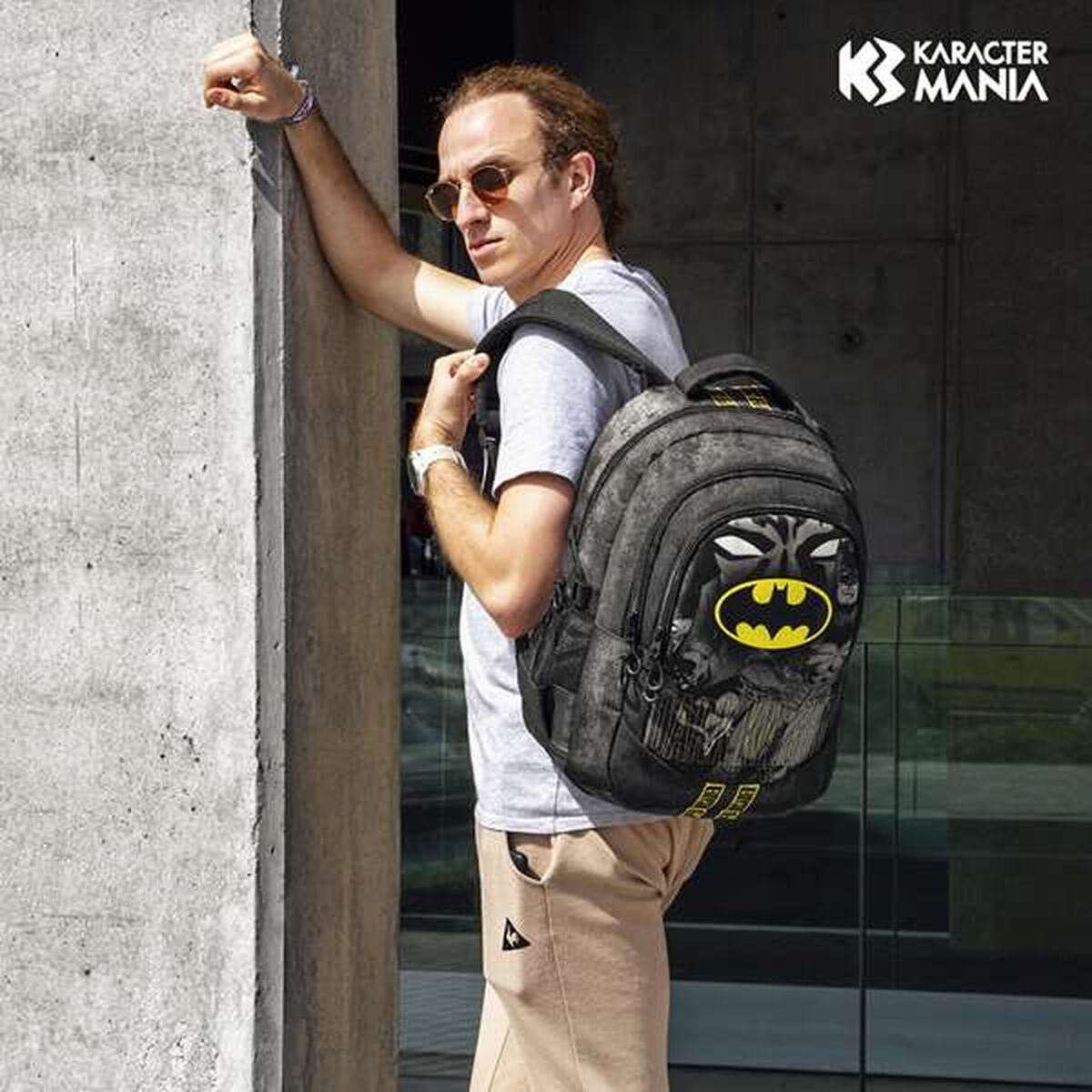 Ein Mann mit Sonnenbrille und Rucksack mit Batman-Logo lehnt an einer Wand.