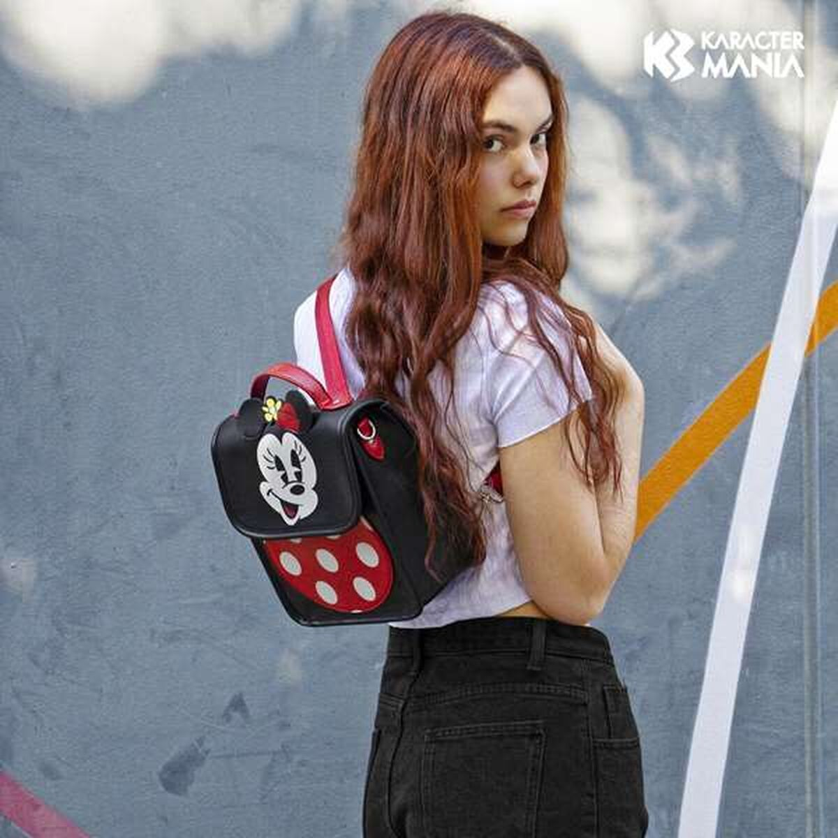 Eine Frau mit roten Haaren trägt einen Minnie Mouse Rucksack. Sie blickt in die Kamera und steht vor einer Wand mit bunten Streifen.