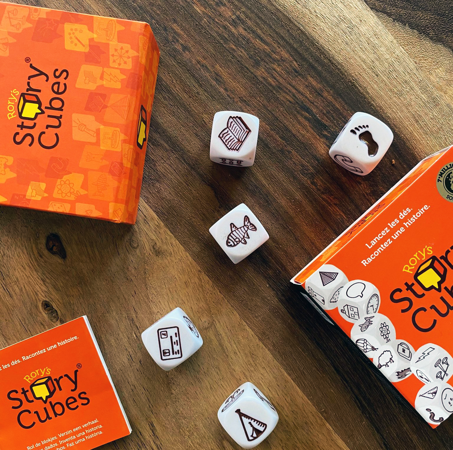 Holztisch mit Rory's Story Cubes Spiel. Würfel mit Symbolen, verstreut um orangefarbene Boxen und ein Heft.