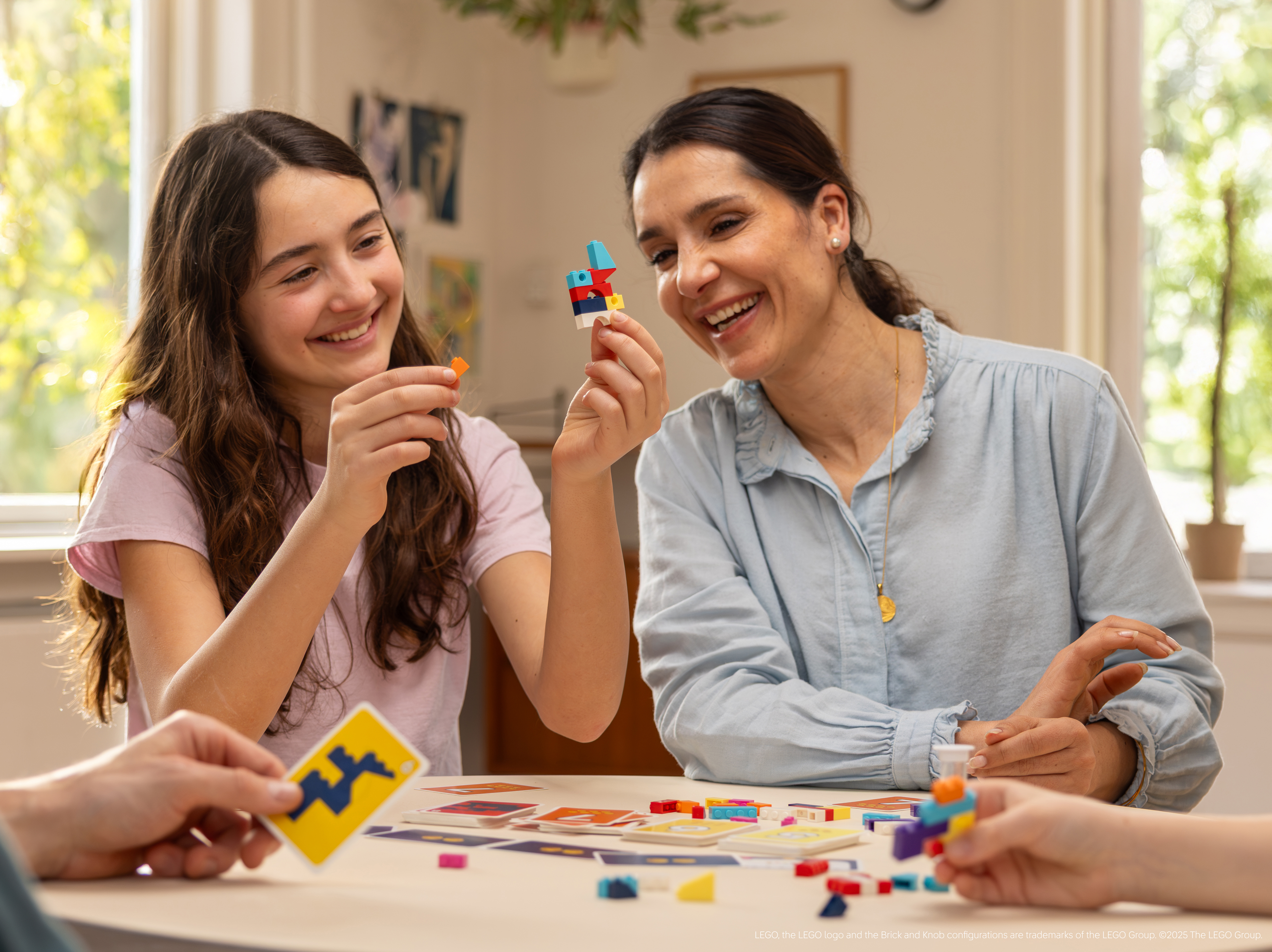 Twee vrouwen en een meisje lachen, spelend met kleurrijke Lego blokken. Een kaart wordt op de voorgrond gehouden.