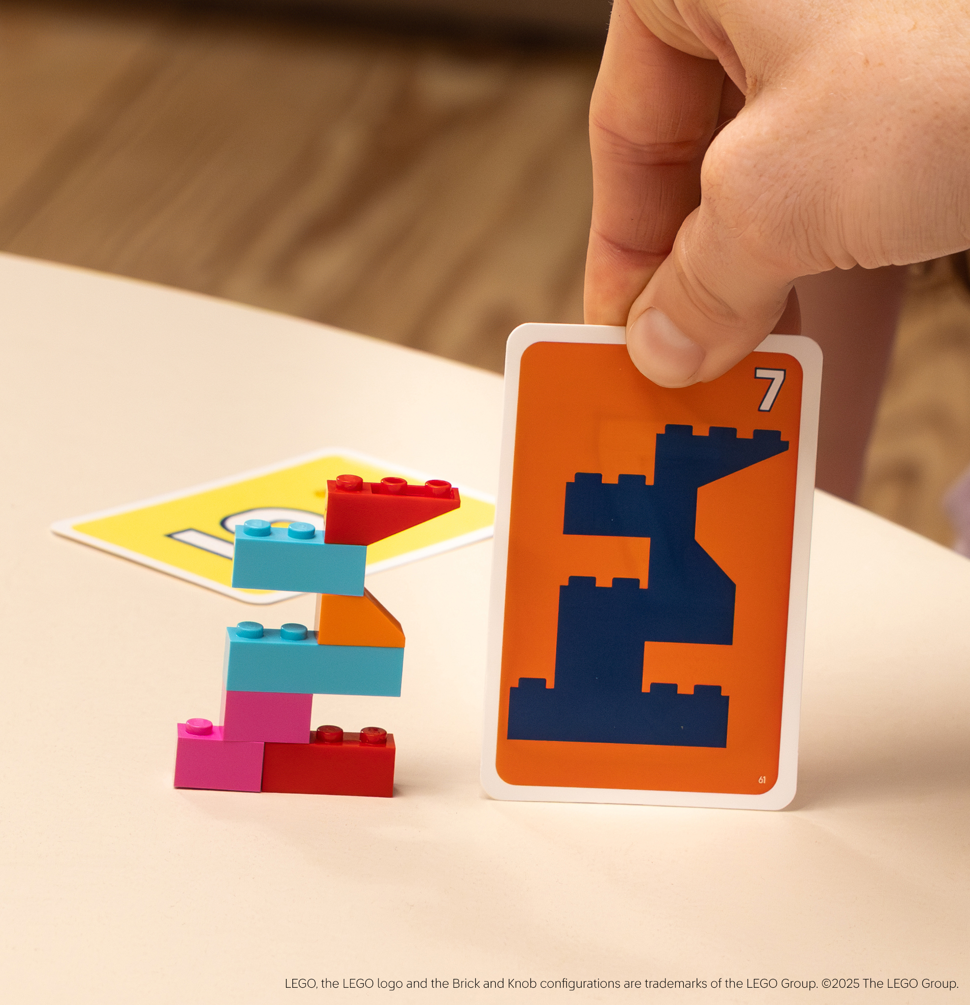 Een hand houdt een kaart met een 7 vast. Legoblokjes vormen een 3. Een andere kaart met een 10 staat op de achtergrond.