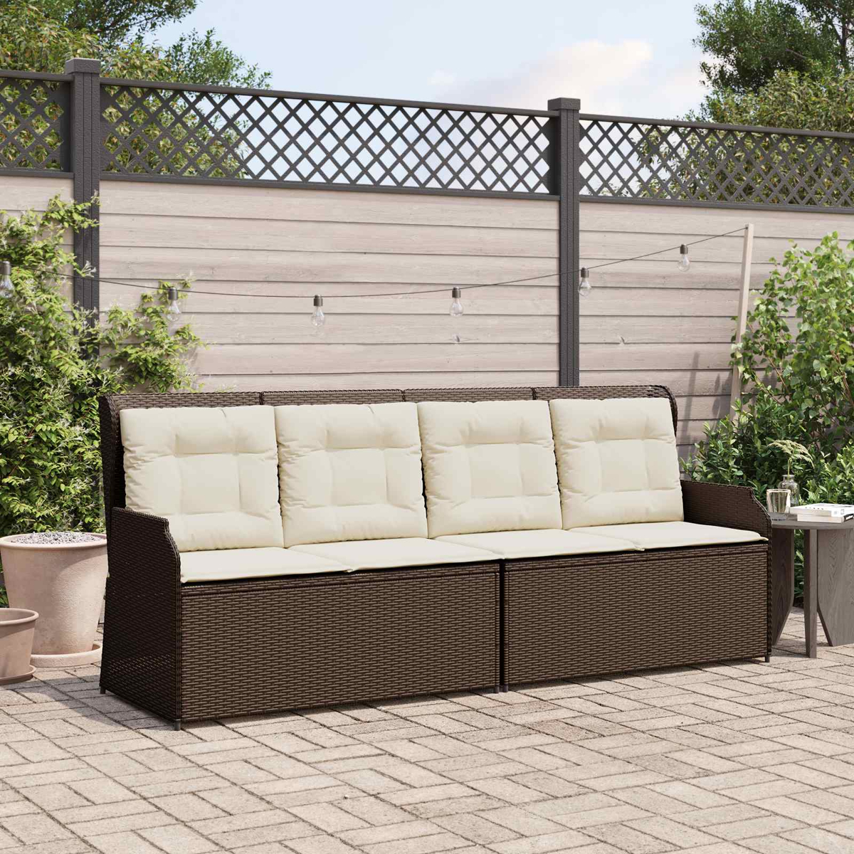 Ein braunes Korbmöbelsofa mit cremefarbenen Kissen steht auf einer Terrasse. Im Hintergrund ist ein Holzzaun mit Gitter oben.