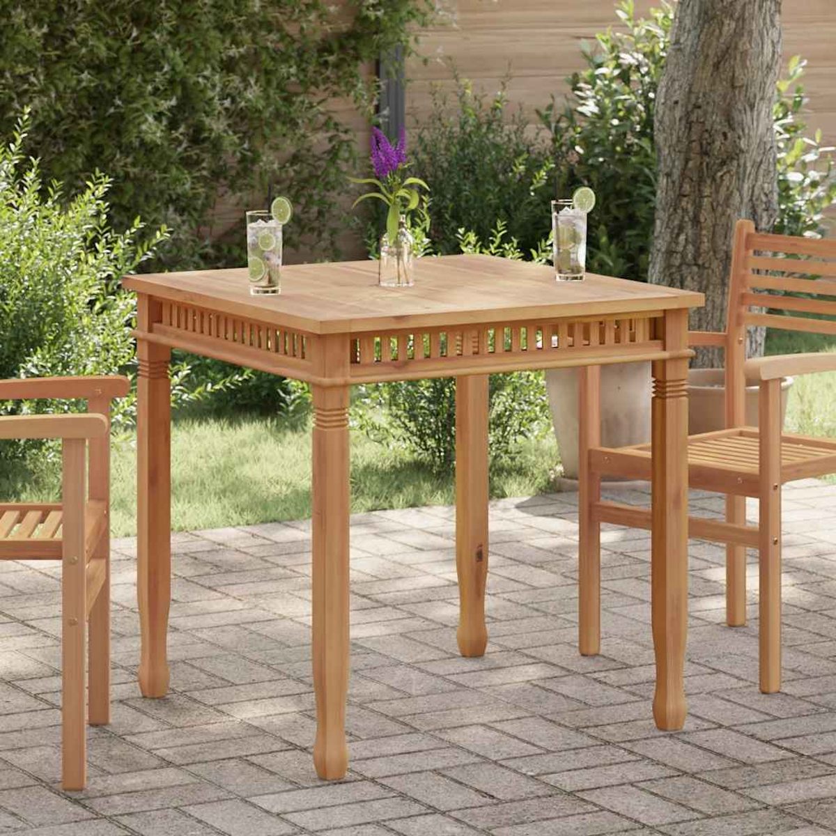 Holztisch mit Getränken, Blumen und Stühlen in einem Garten. Im Hintergrund Büsche und ein Baum. Pflastersteine sind sichtbar.