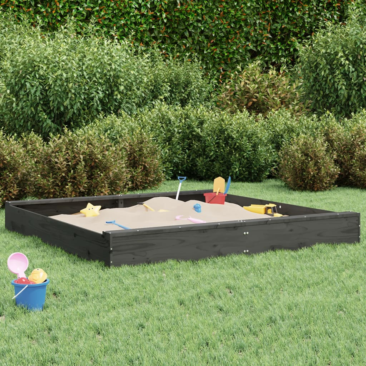 Ein Sandkasten mit Spielzeug steht auf grünem Gras. Büsche bilden den Hintergrund. Ein blauer Eimer mit Spielzeug befindet sich im Vordergrund.
