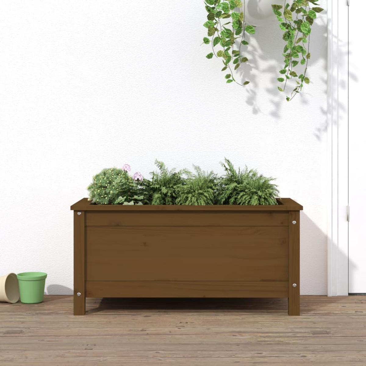 Eine braune Holzkasten mit grünen Pflanzen und kleinen rosa Blumen. Im Hintergrund eine weiße Wand und Tür.