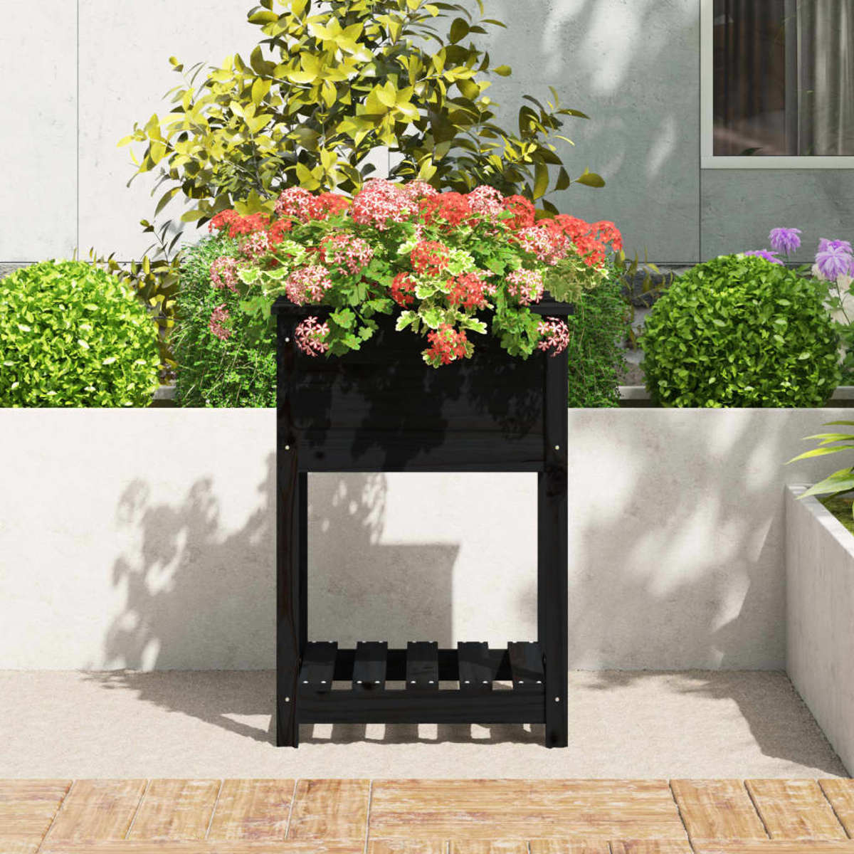 Ein schwarzer Pflanzkasten mit roten Blumen steht auf einer Terrasse, umgeben von grünen Büschen und einem Gebäude im Hintergrund.