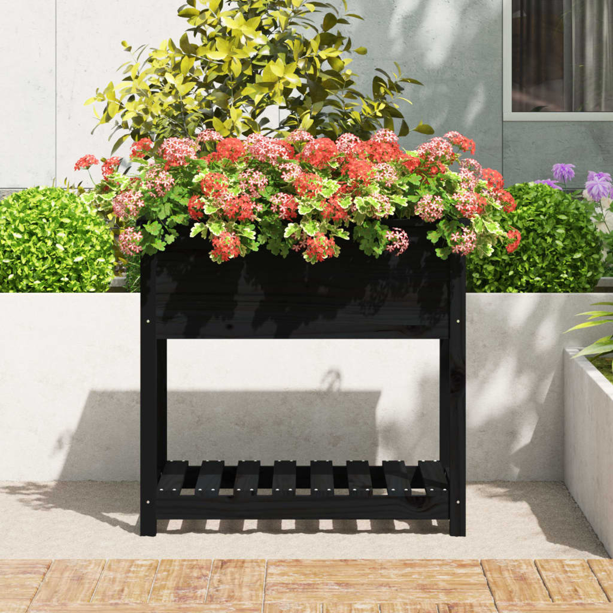 Eine schwarze Holzkiste mit roten und rosa Blumen und grünen Blättern. Der Hintergrund umfasst grüne Büsche und ein Gebäude.