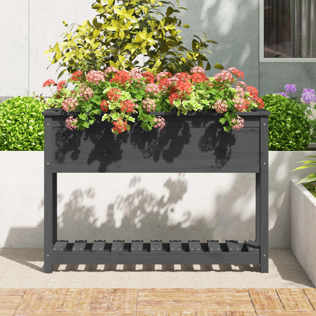 Eine dunkelgraue Holzpflanzkasten mit roten und grünen Blumen. Dahinter eine Betonmauer und Grün. Der Pflanzer steht auf einer Terrasse.