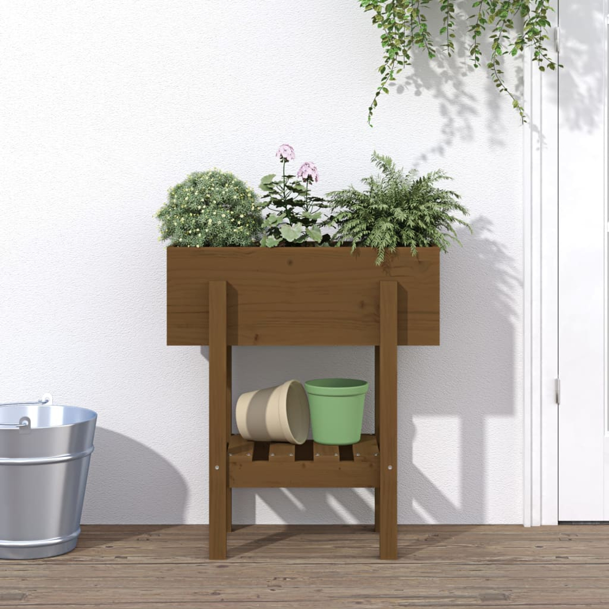 Ein Holzpflanzer mit Pflanzen steht auf einem Deck. Zwei Töpfe und ein Eimer sind ebenfalls vorhanden. Der Hintergrund ist eine weiße Wand und eine Tür.