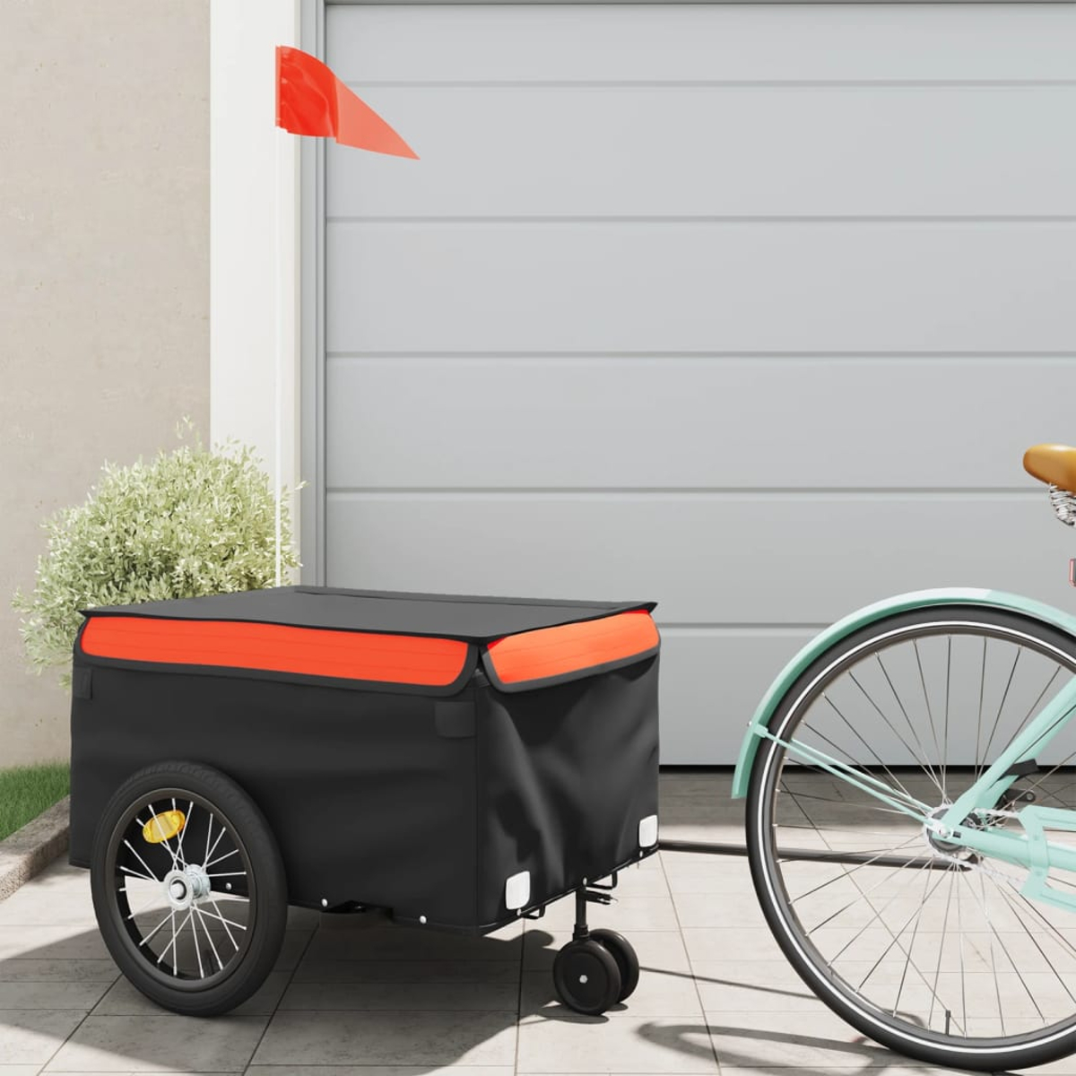 Ein Fahrradanhänger mit schwarzem Körper und orangefarbener Verkleidung. Eine rote Fahne ist befestigt. Daneben steht ein hellblaues Fahrrad, im Hintergrund die Garage.