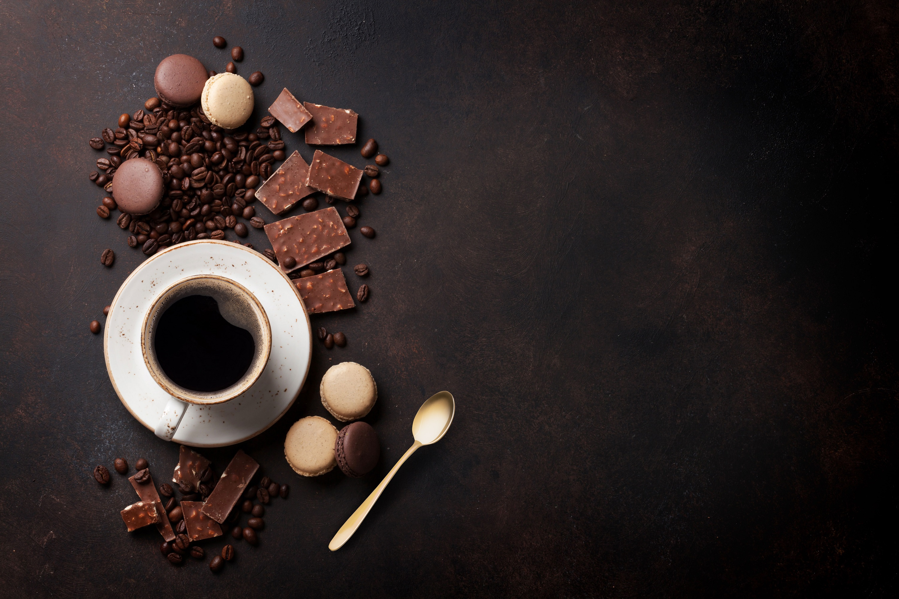Overhead shot of coffee cup, macarons, chocolate and coffee beans on dark brown surface.